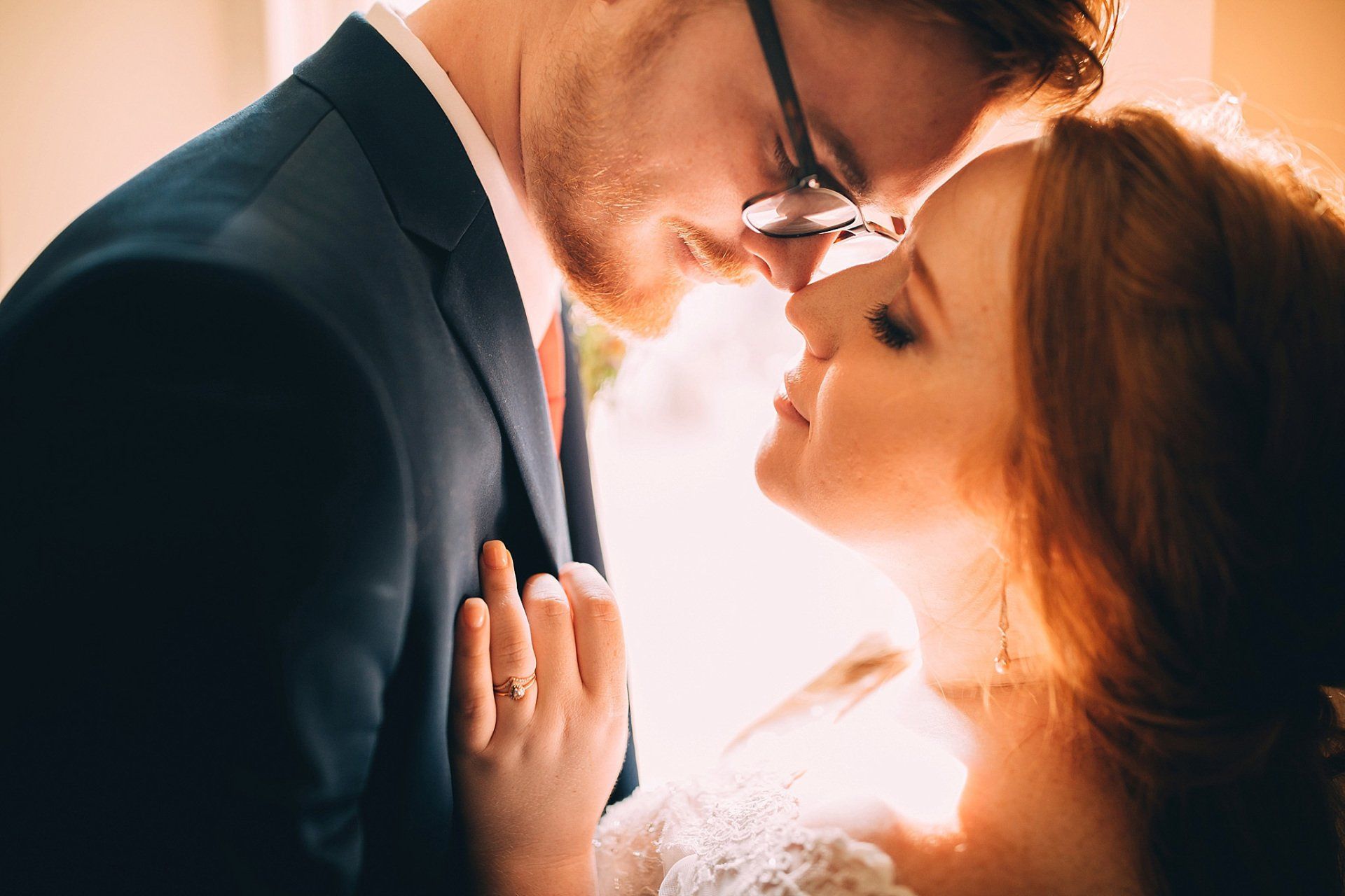 Bride and groom embrace, foreheads touching, sunlight. She wears white, he a blue suit, both smiling.