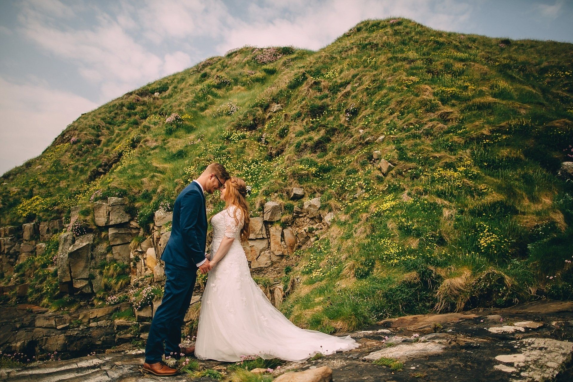 Couple in wedding attire facing each other, head to head, in front of a green hillside.