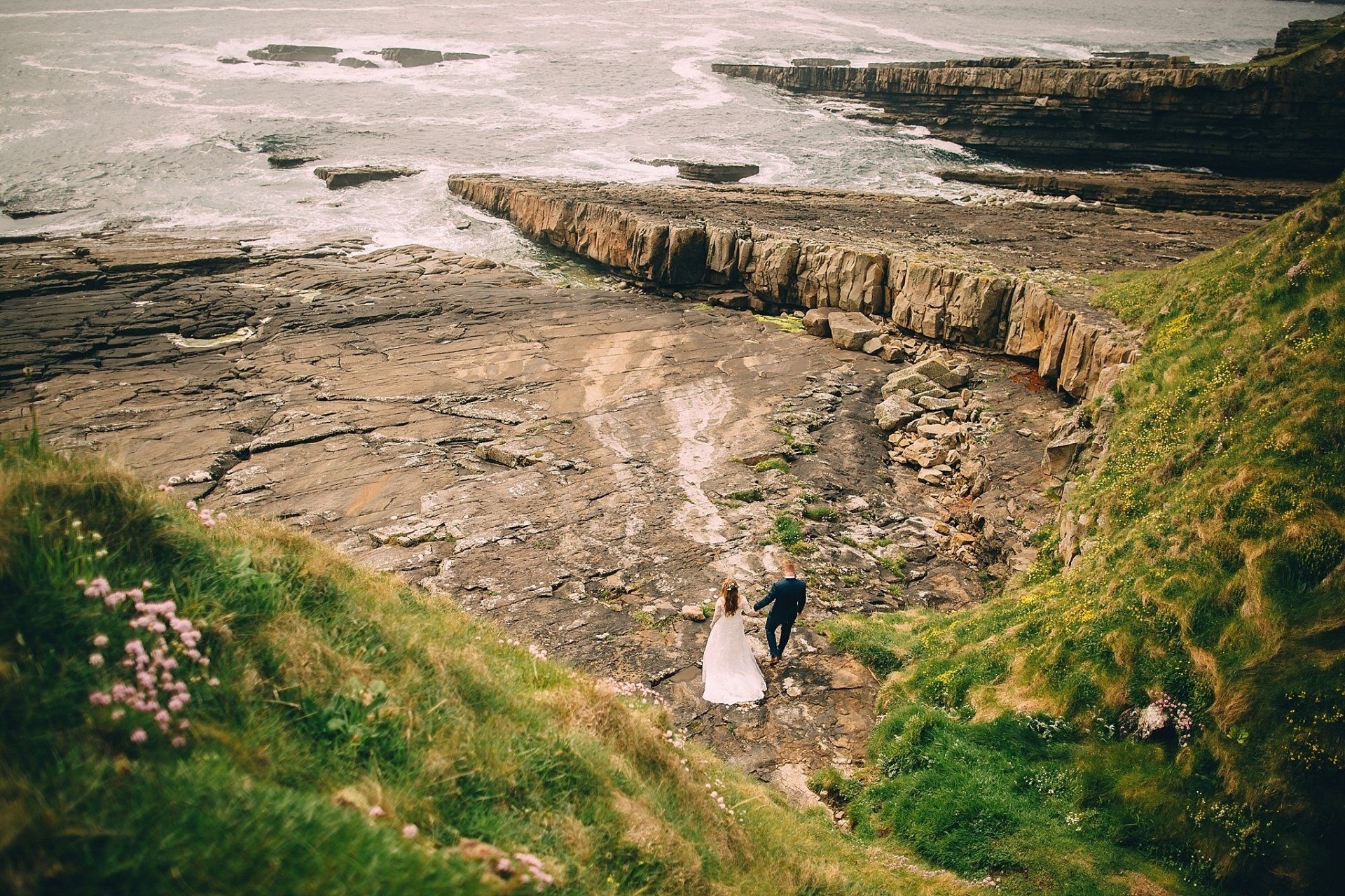 Couple in wedding attire walk on rocky shore near ocean. Green grass and flowers in foreground.