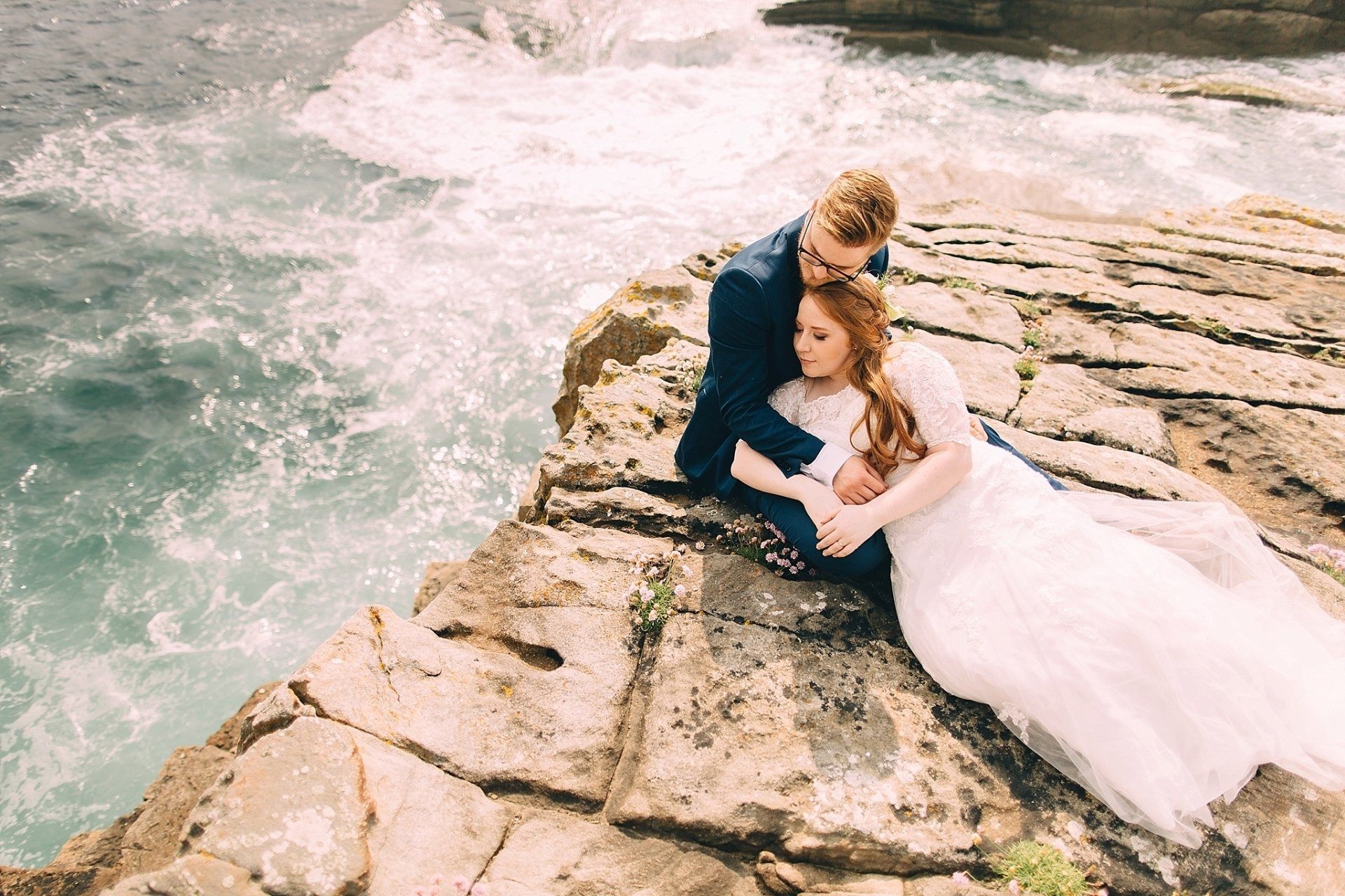 Couple in wedding attire embrace on a rocky cliff, ocean waves crashing nearby.