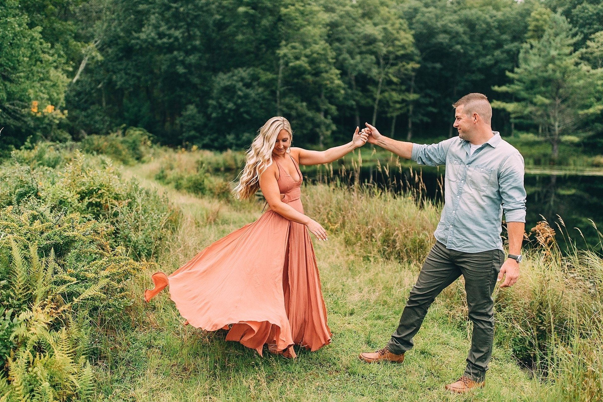 Couple dancing outdoors by a pond; woman in flowing orange dress, man in denim shirt and camo pants.
