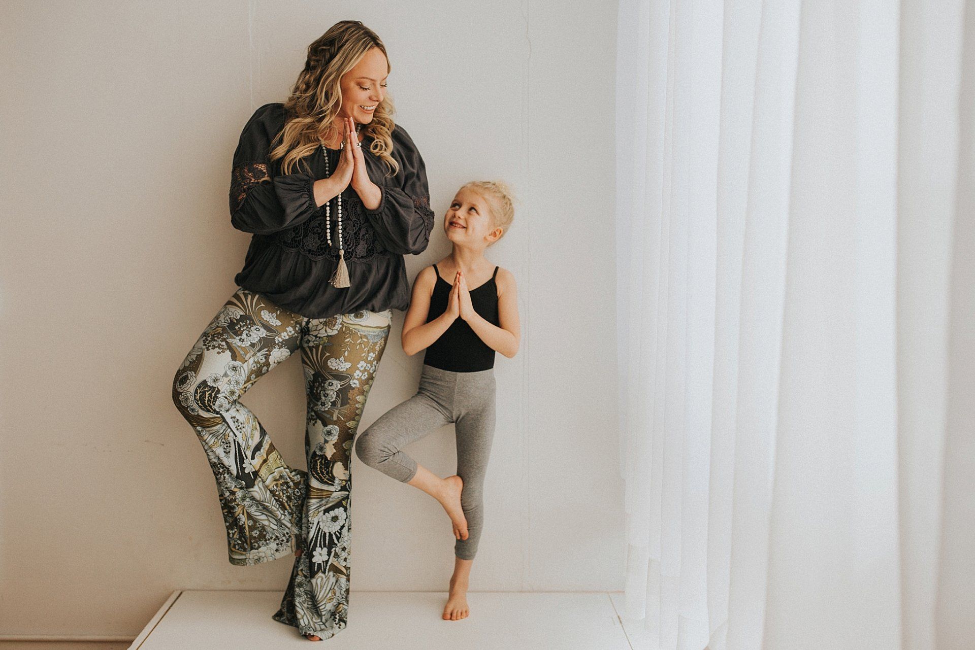 Woman and child in yoga pose, hands together, smiling, near a white wall and curtain.