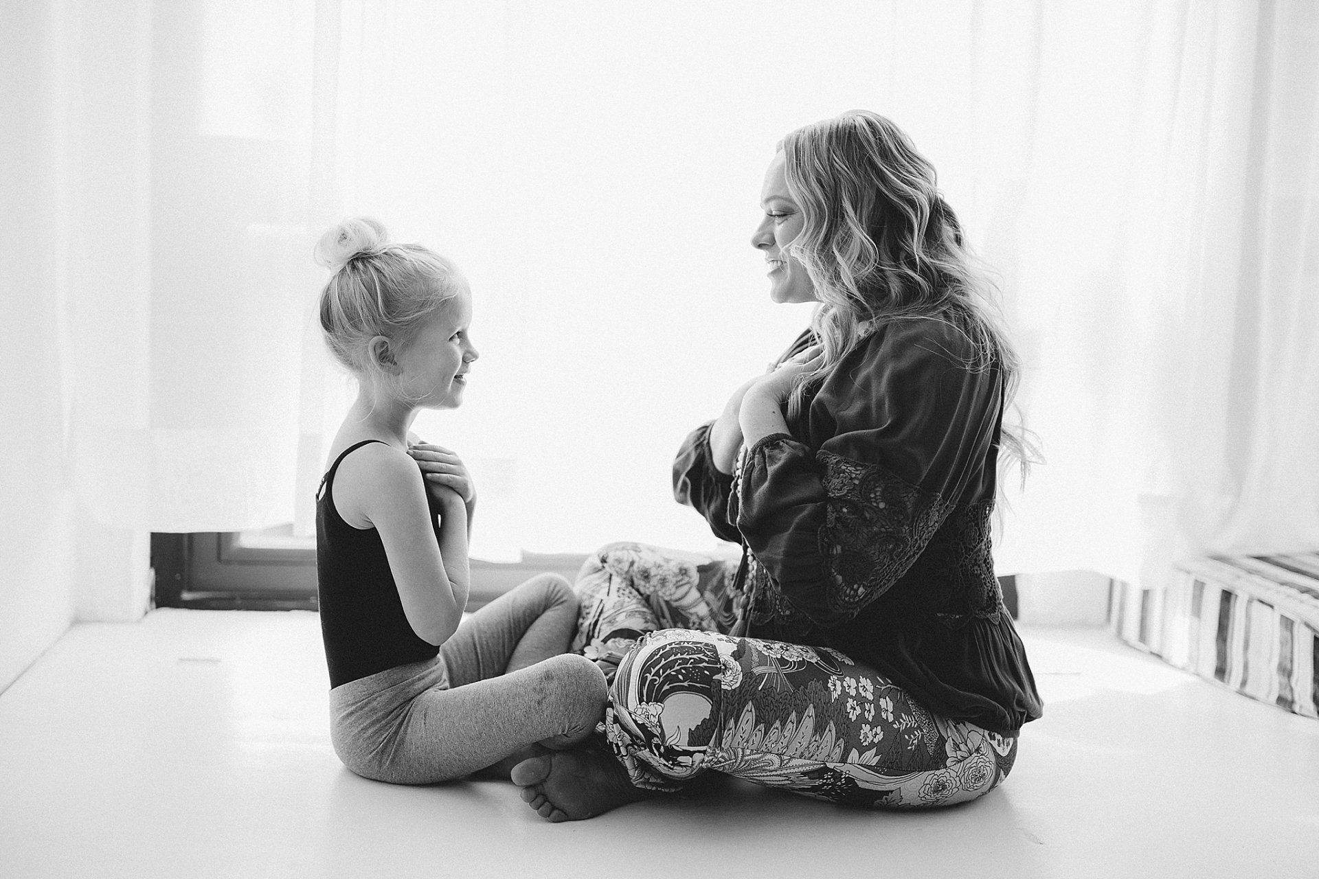 Woman and young girl seated on the floor, smiling and holding hands over their hearts, indoors.