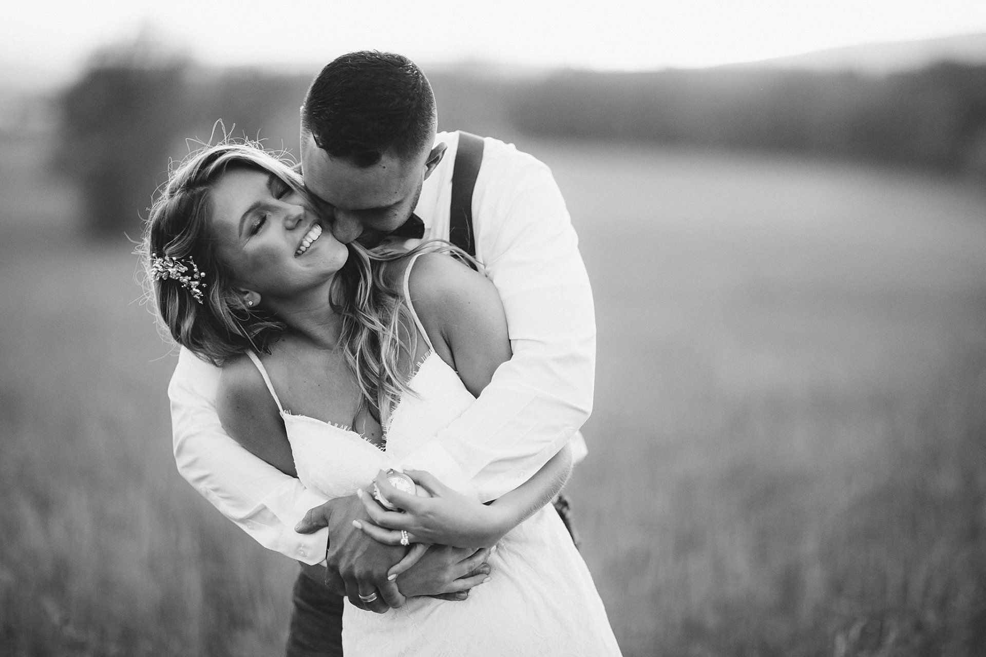 Couple embracing, woman laughing, man kissing her neck, outdoor field, black and white.