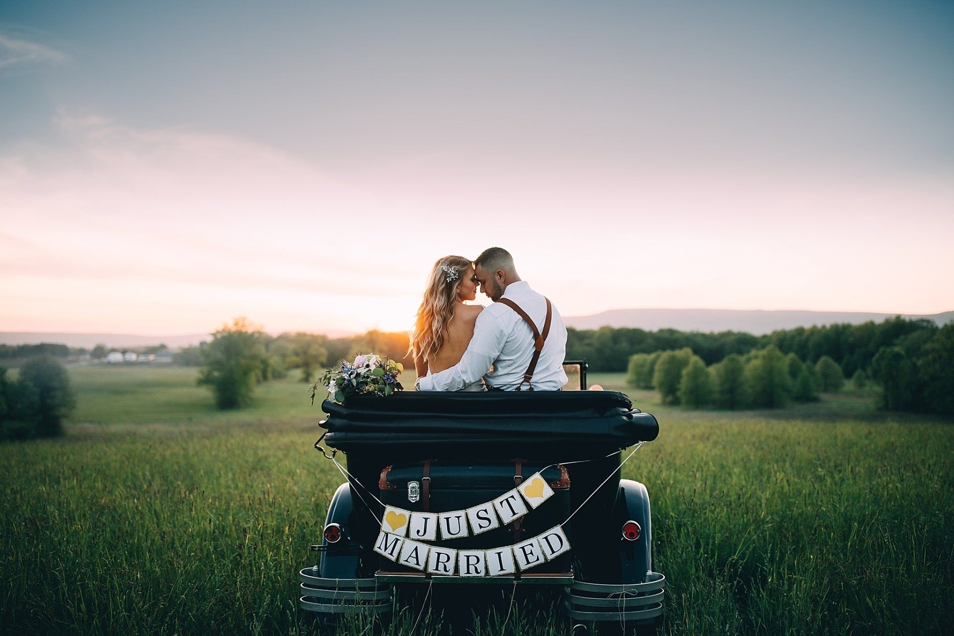 Newlyweds in antique car, heads touching, field at sunset.