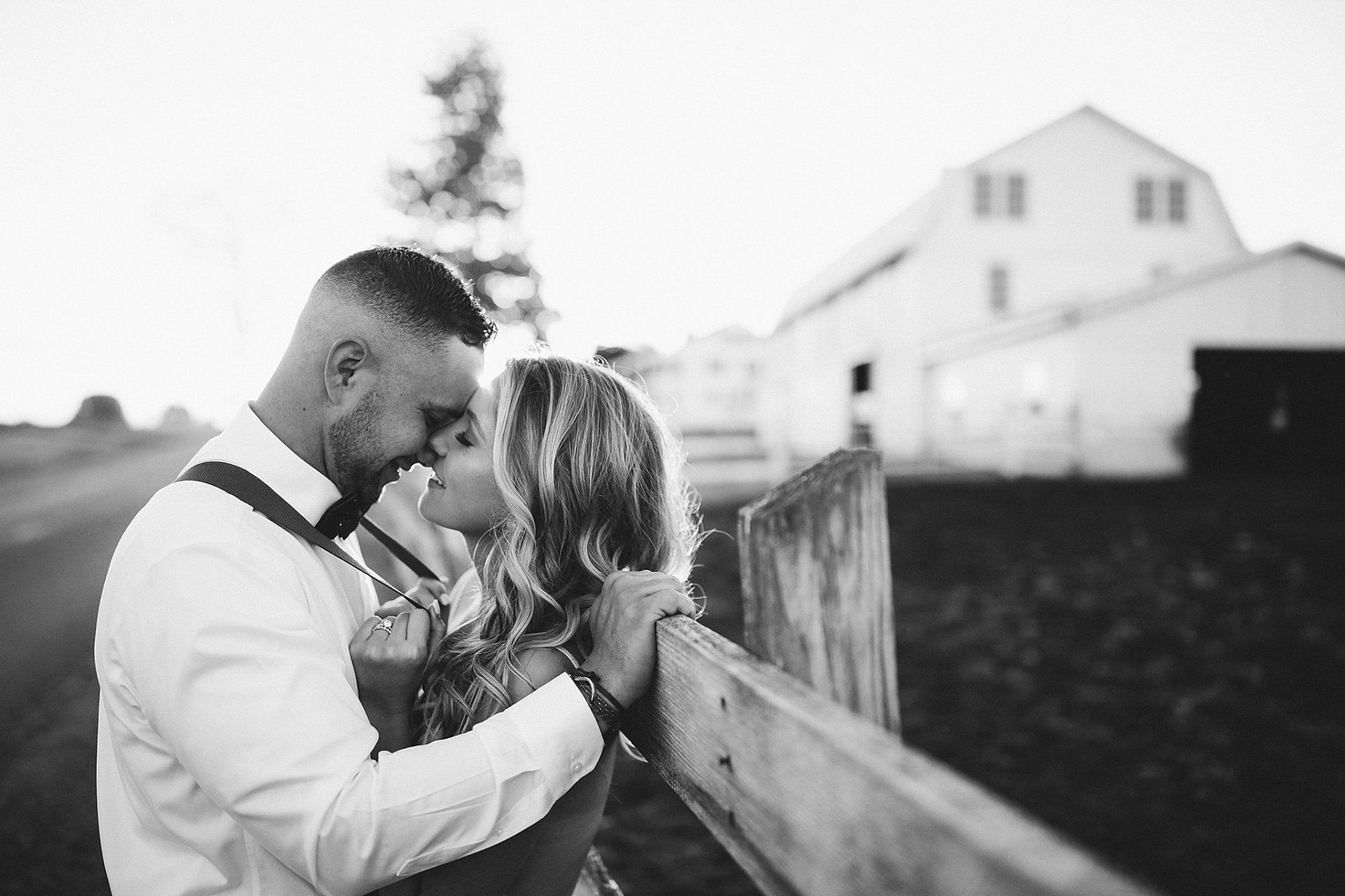 Couple embraces near a wooden fence, with a barn in the background. Black and white.