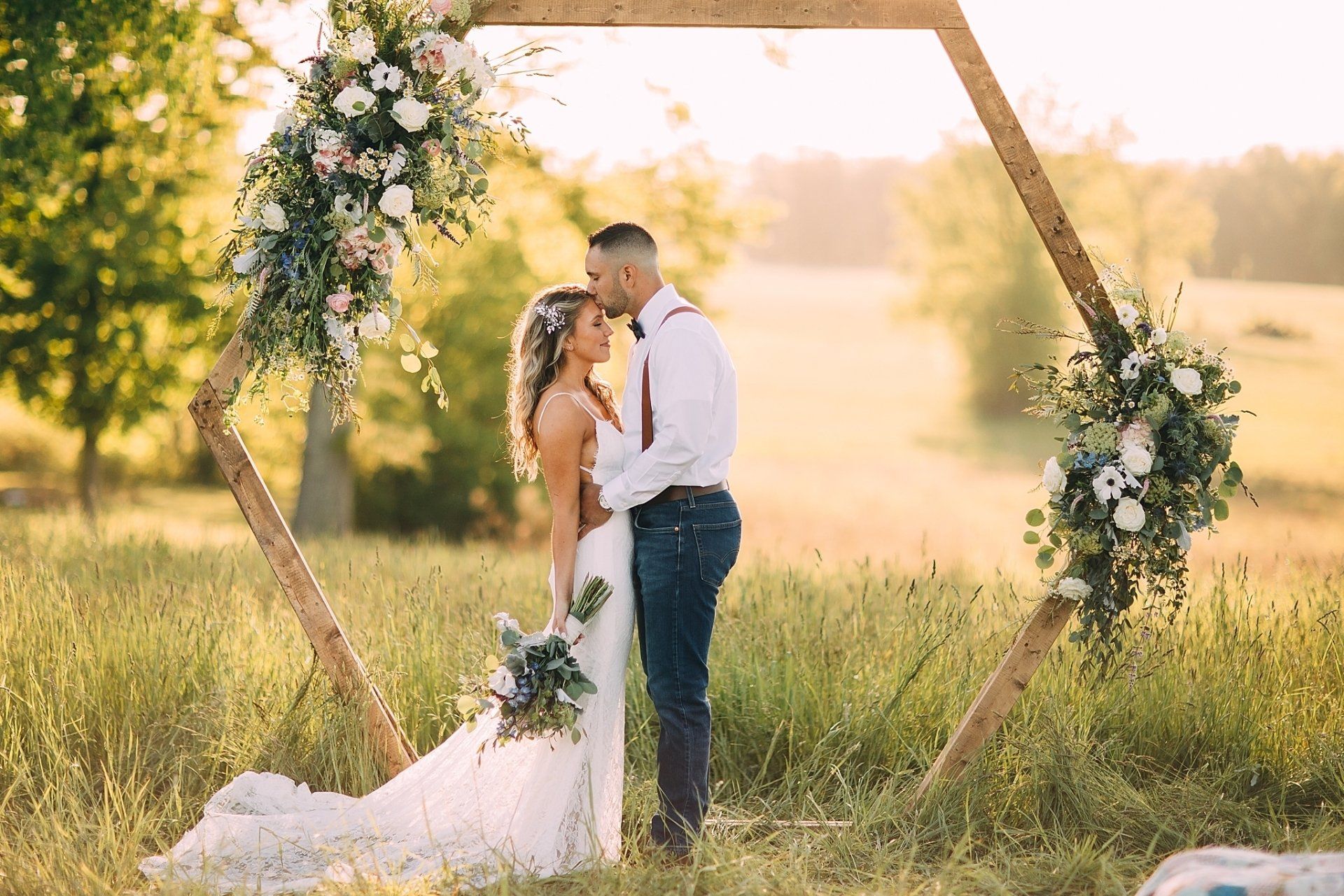 Couple embraces under a flower-decorated hexagon arch in a field; the man kisses the woman's forehead.
