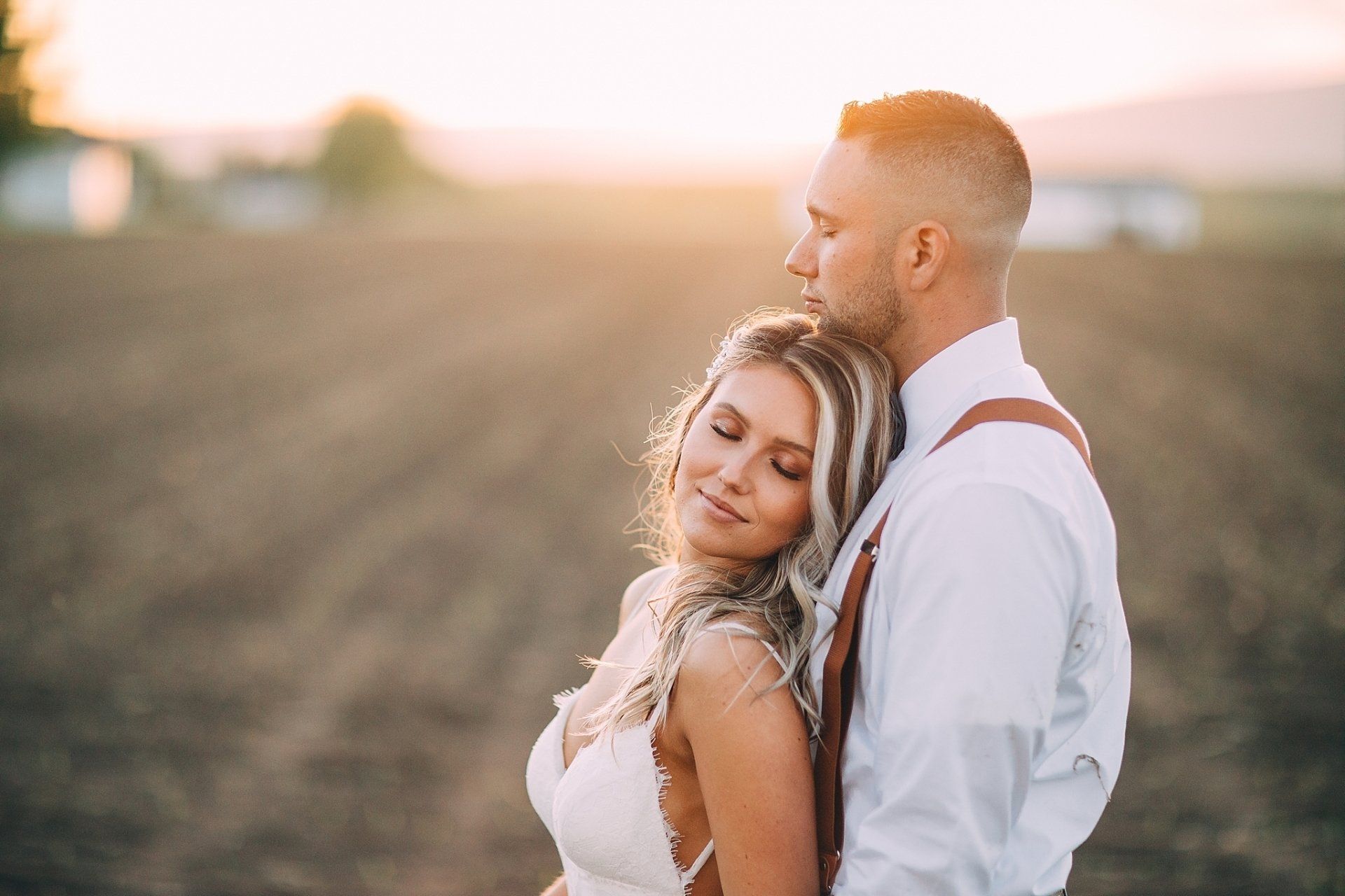 Couple embracing in a field at sunset; woman in white dress, man in white shirt and suspenders.