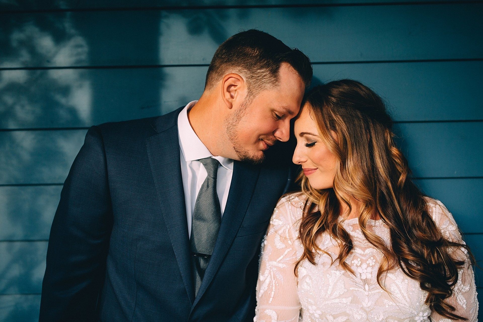 Couple in wedding attire leaning heads together against blue wall.
