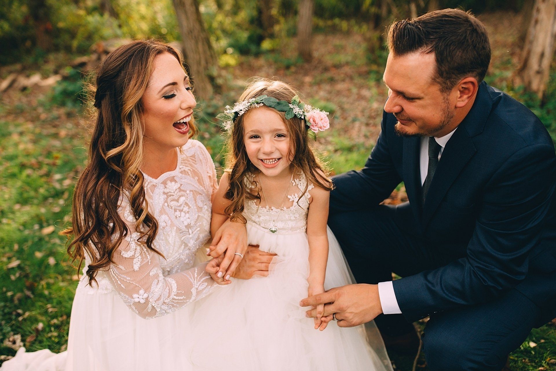 Newlyweds and flower girl laughing in a field. Bride in white lace, groom in a navy suit, flower girl in a floral crown.