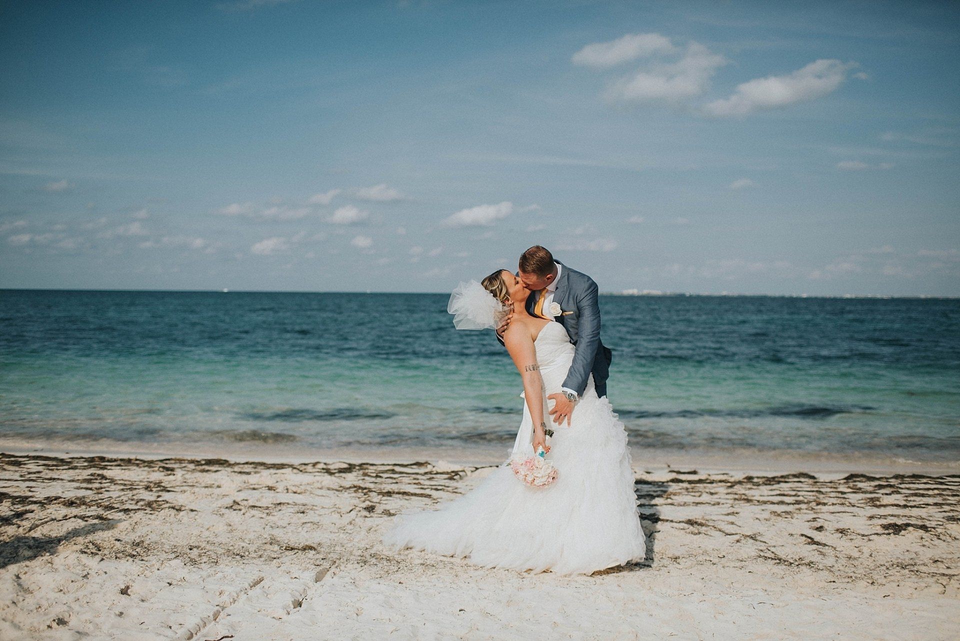Couple kissing on a beach. Bride in white dress, groom in blue suit. Ocean and sky visible.