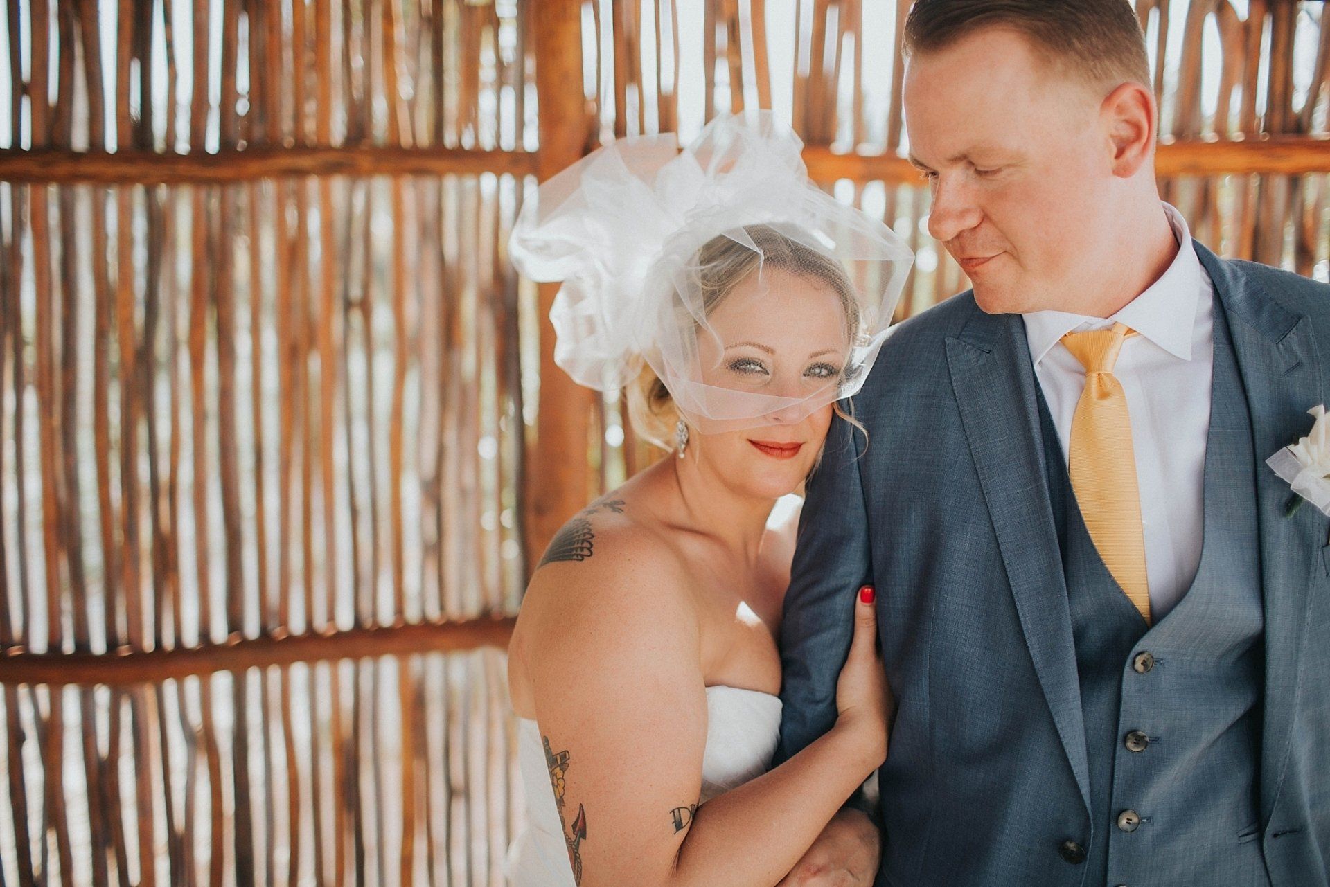 Bride and groom in wedding attire pose by woven wooden wall. Bride in veil, groom in suit.