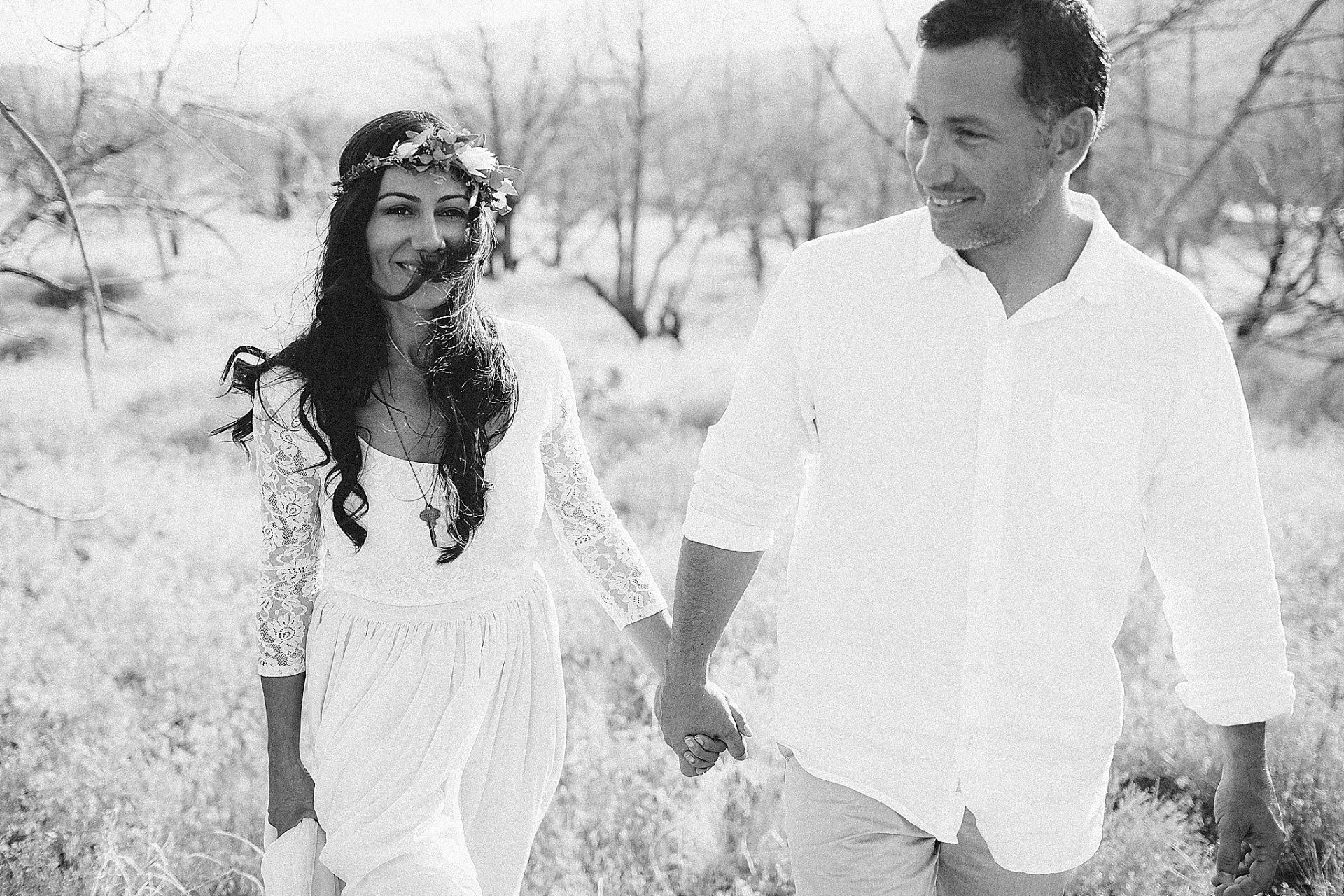 Couple holding hands, walking through a field; woman in dress with floral crown, man in white shirt.