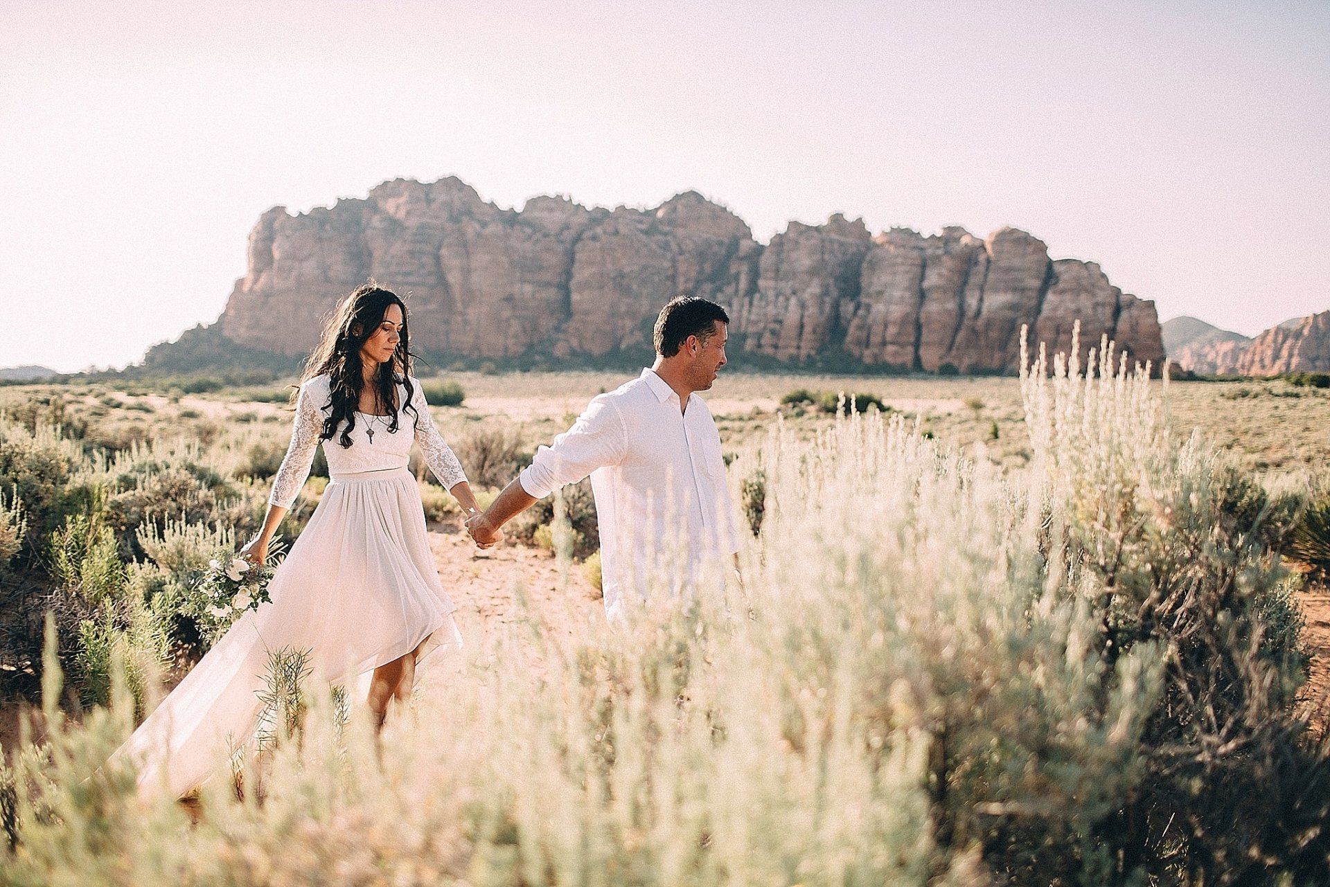 Couple holding hands, walking through desert landscape toward rock formation. Soft light and neutral colors.