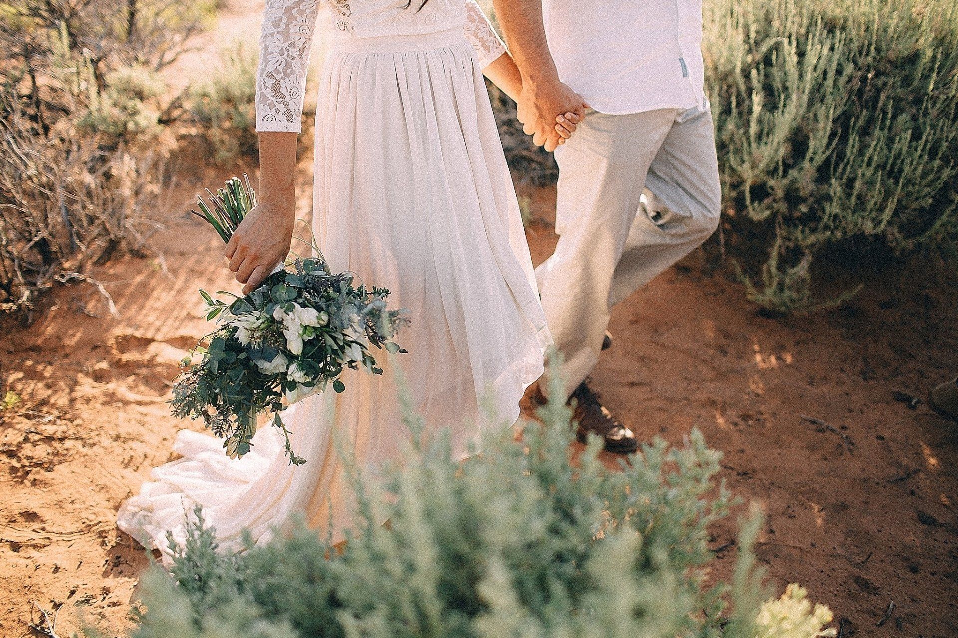Couple holding hands, bride in white dress holding bouquet, walking in desert setting.
