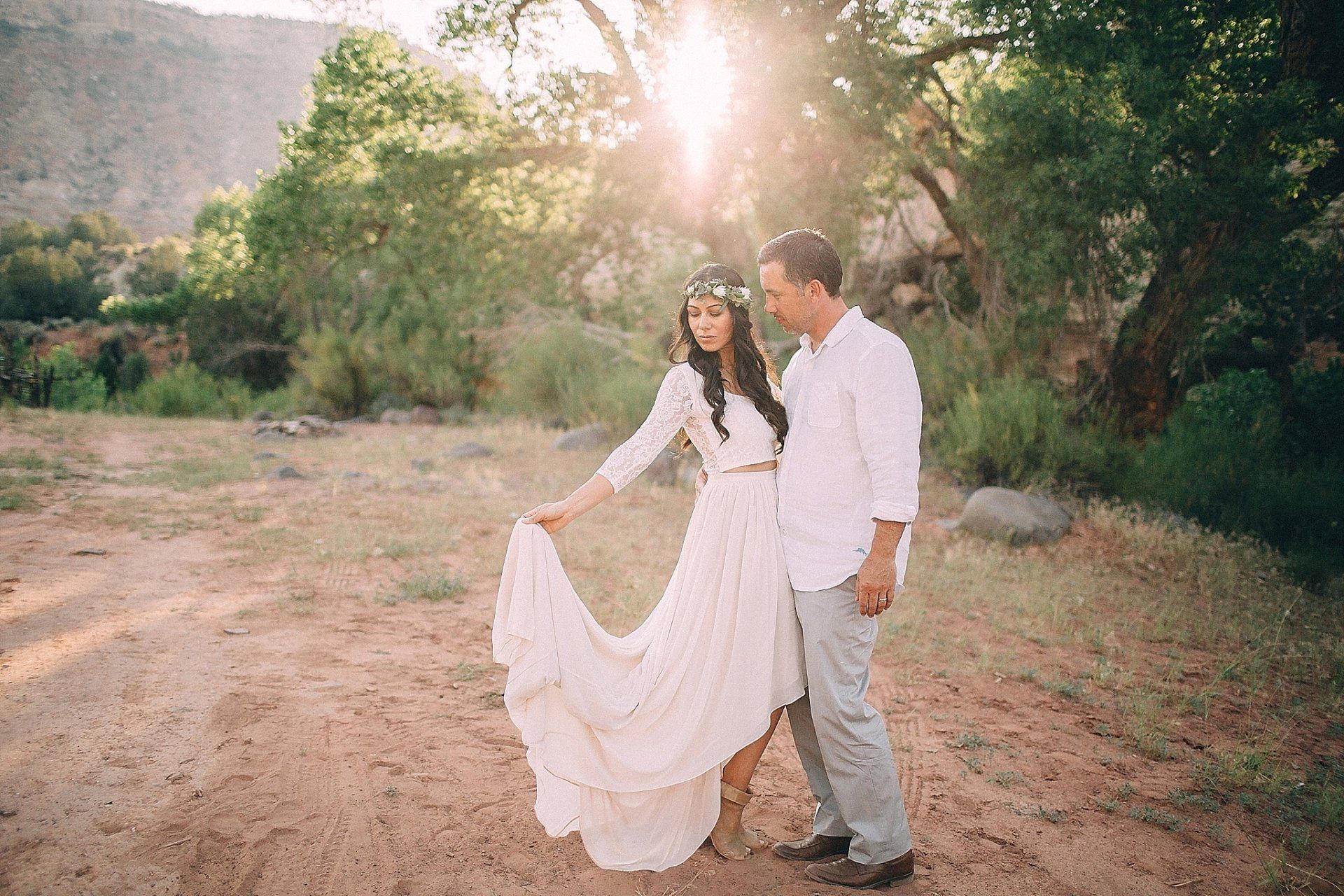 Couple poses outdoors in sunlight; woman in white dress, flower crown, man in white shirt, trees in background.