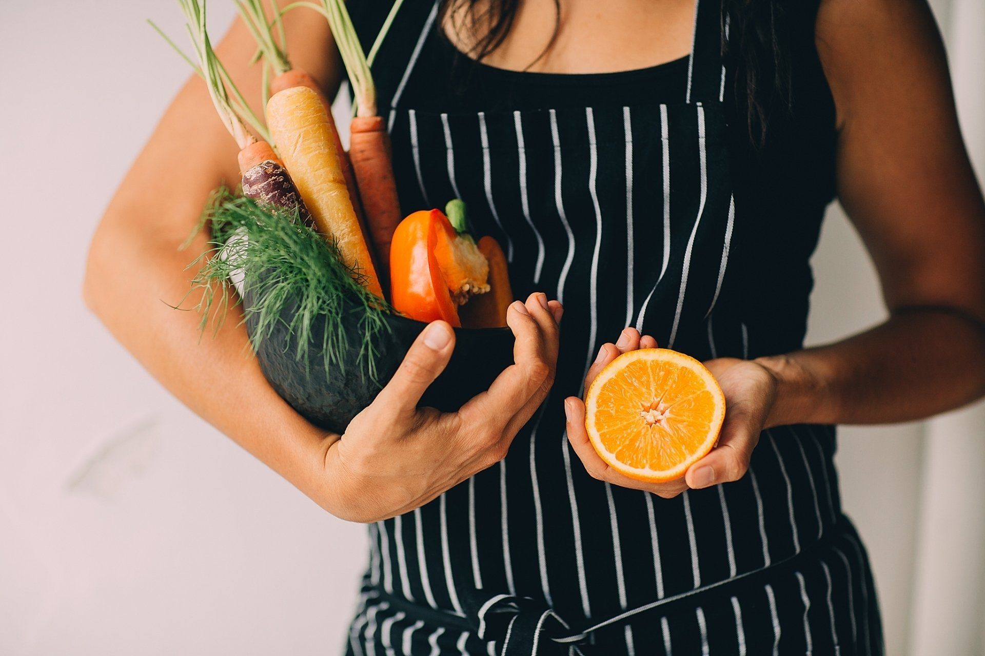 Woman in striped apron holds vegetables and orange half.