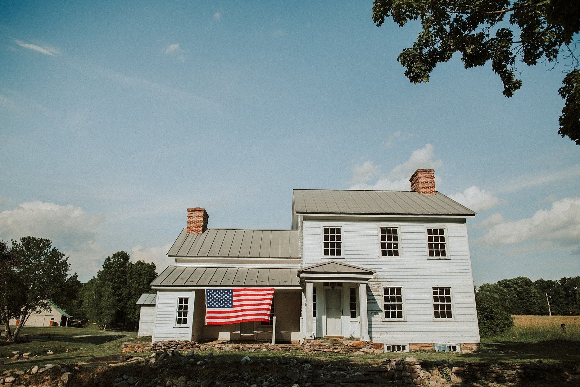 Two-story white house with American flag hanging on the porch. Blue sky and trees in the background.