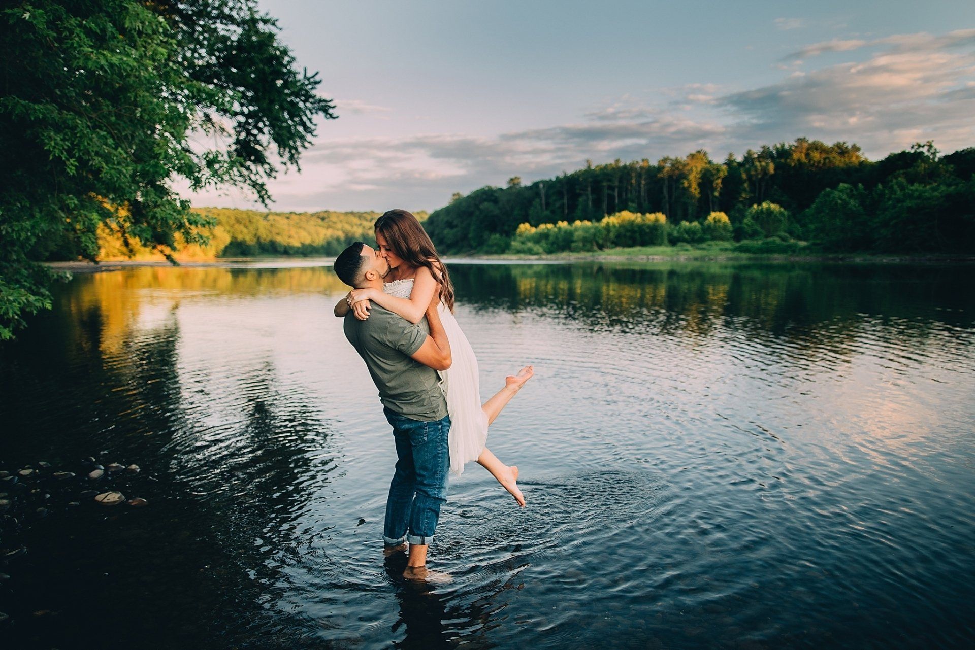 Couple kissing in water; man holding woman up, lake and trees in background.