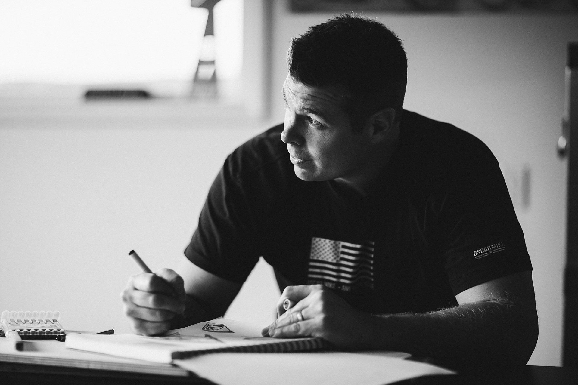 Man in black shirt writing in notebook, looking off to the side, indoors.