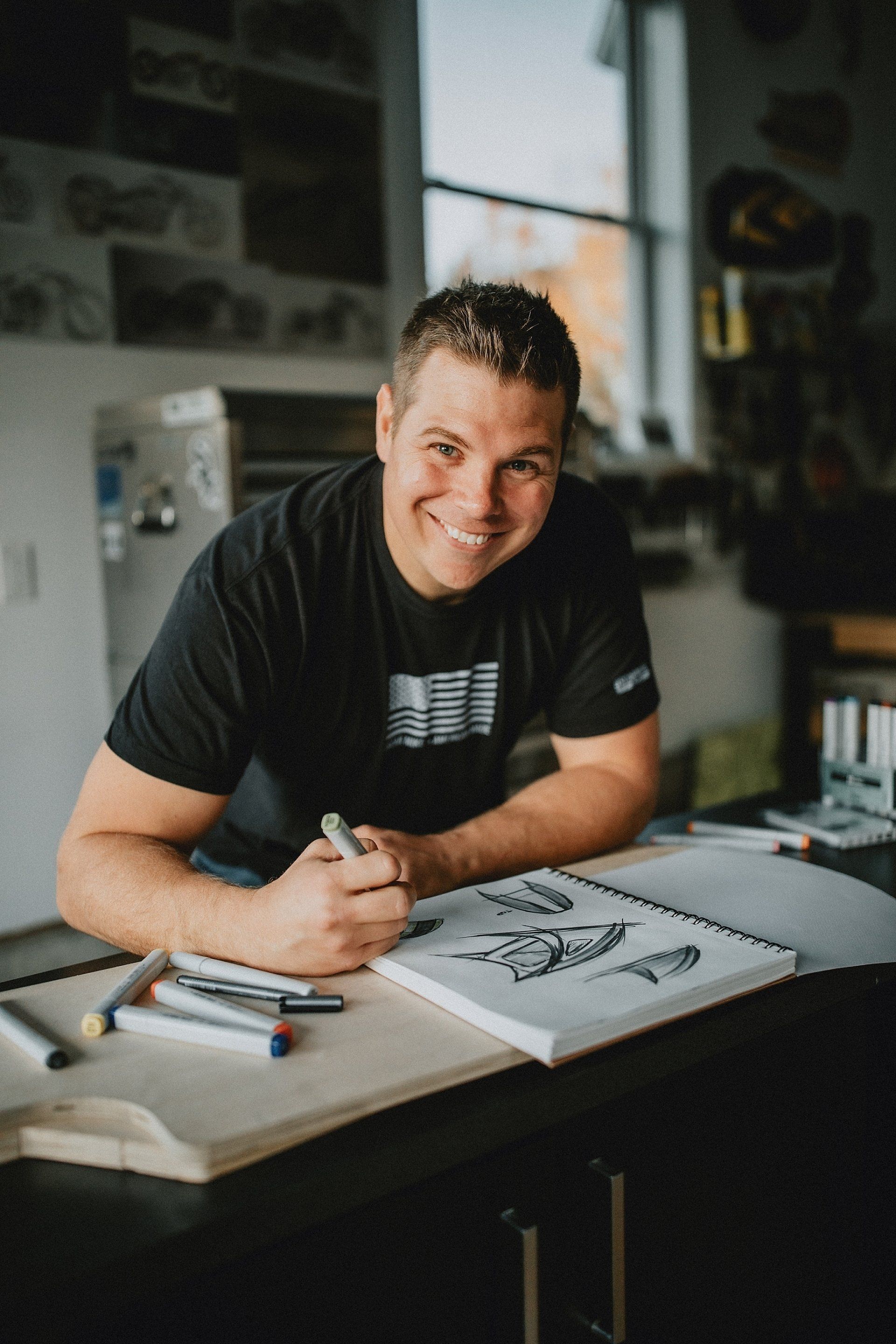 Man smiling, drawing in a notebook with markers on a table.