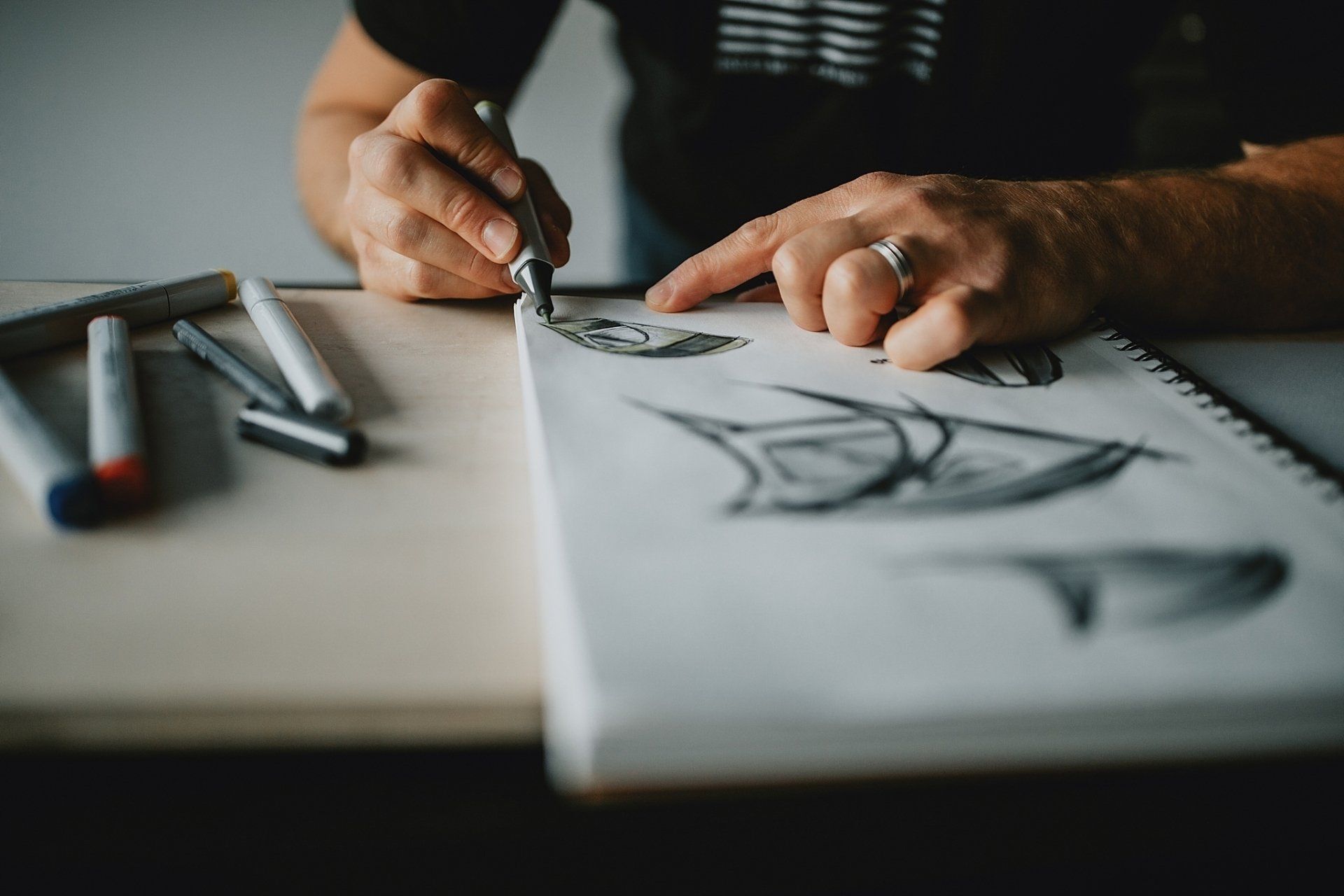 Person sketching on a white pad with a dark pen and pointing finger, art supplies on desk.