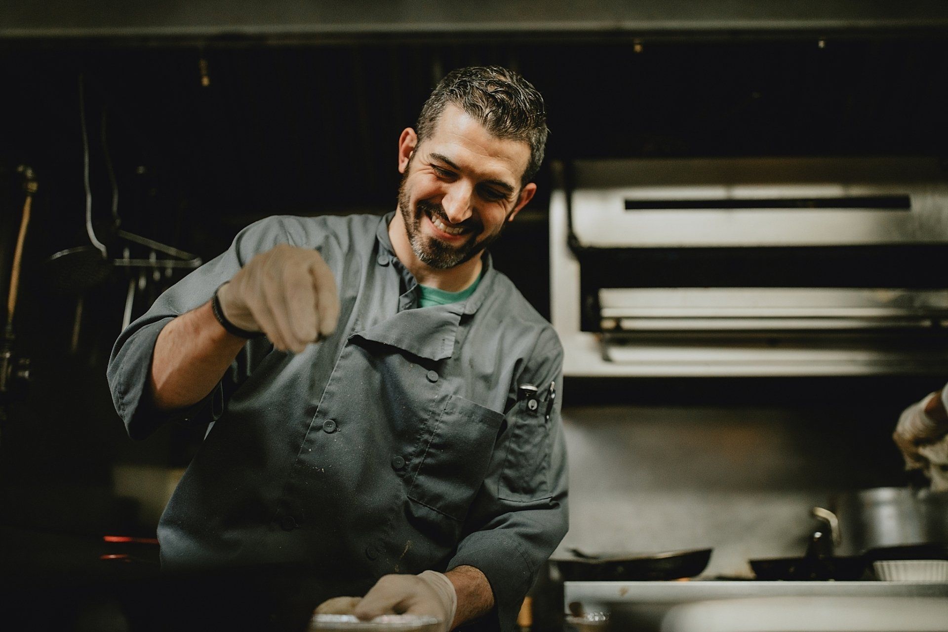 Chef in gray uniform, smiling, sprinkling seasoning over food in a kitchen.
