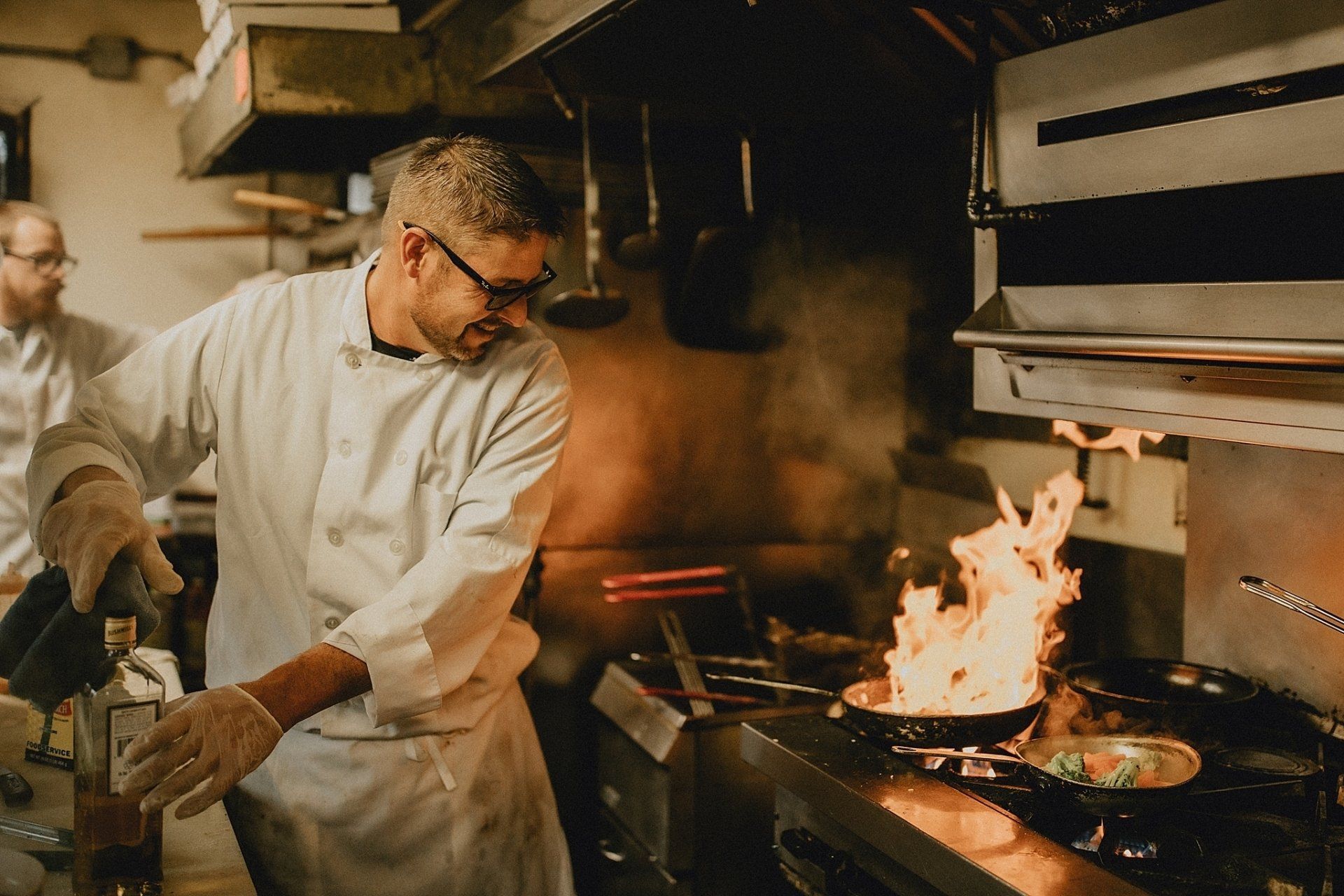 Chef in white coat, glasses, and gloves flambés food in pan on a stove in a kitchen.