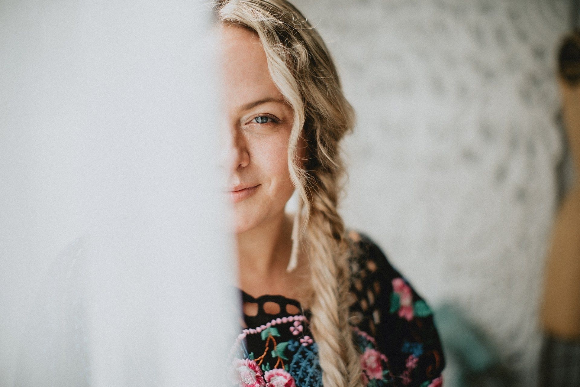 Blonde woman peeks from behind white curtain, smiling. Wearing a floral top with a braid.
