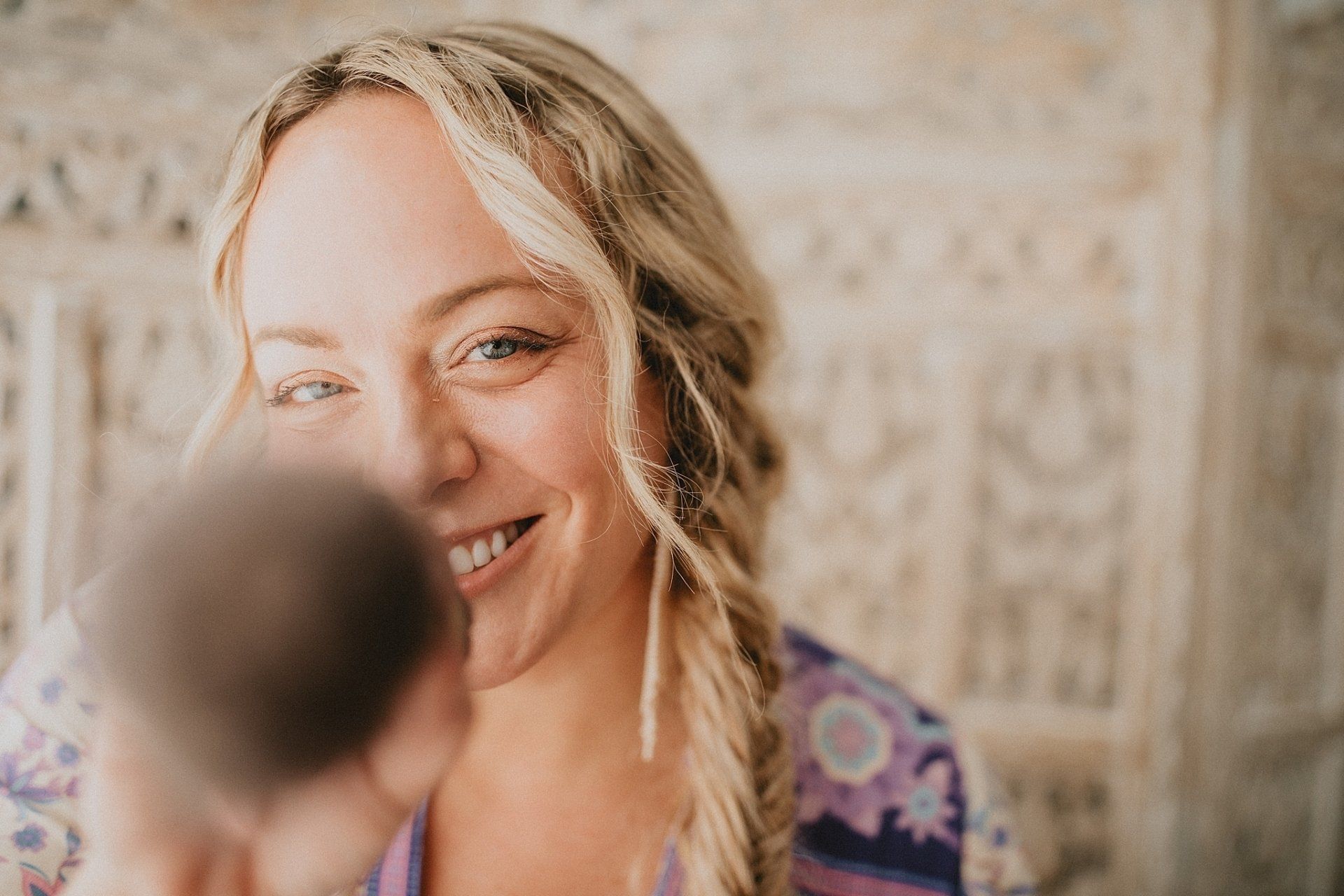 Blonde woman smiles, holding blurred object, against a carved, light-colored wooden screen.