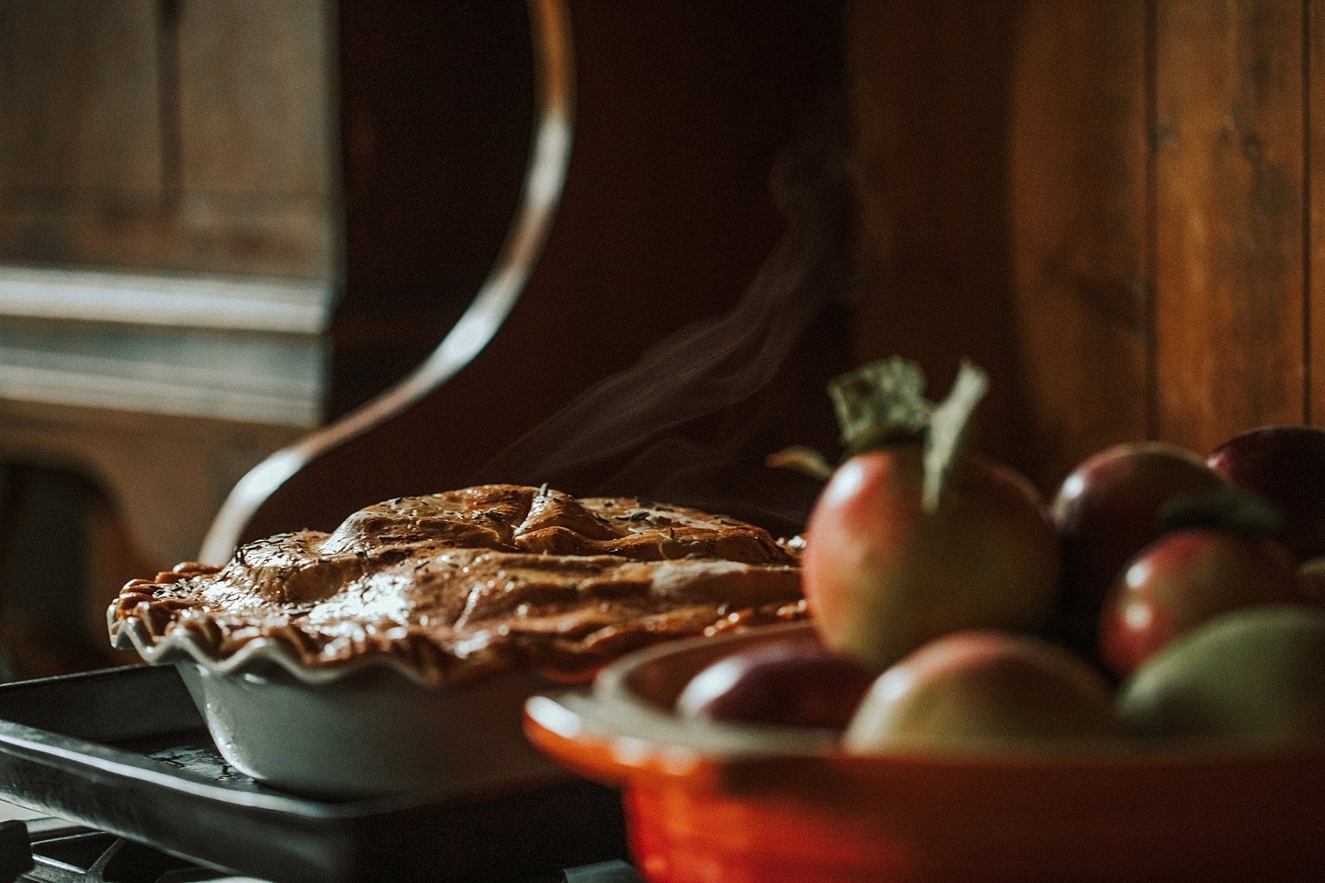 Apple pie in white dish next to bowl of apples on a countertop, dimly lit kitchen setting.