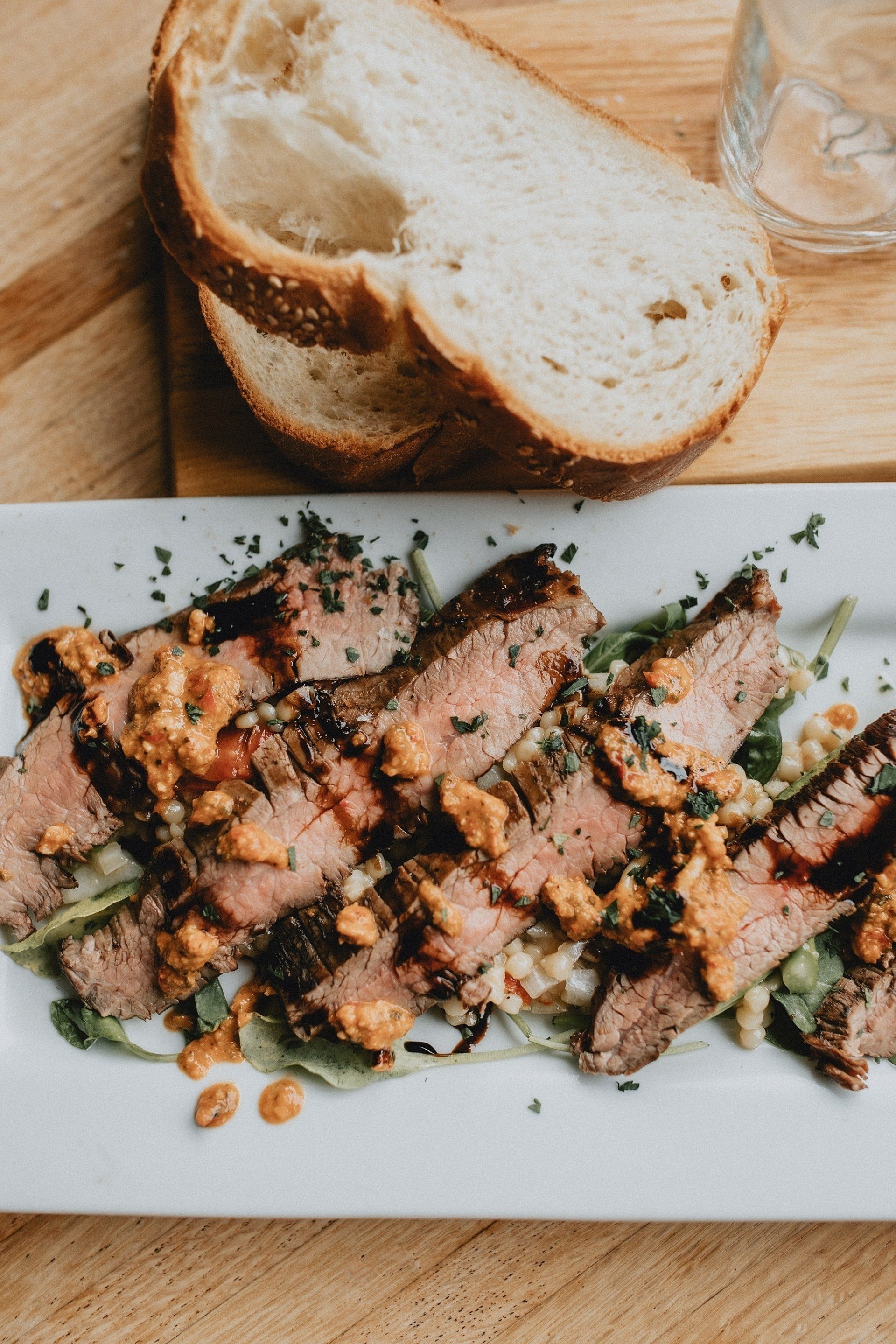 Sliced steak with sauce on a bed of greens, served with bread on a white platter.