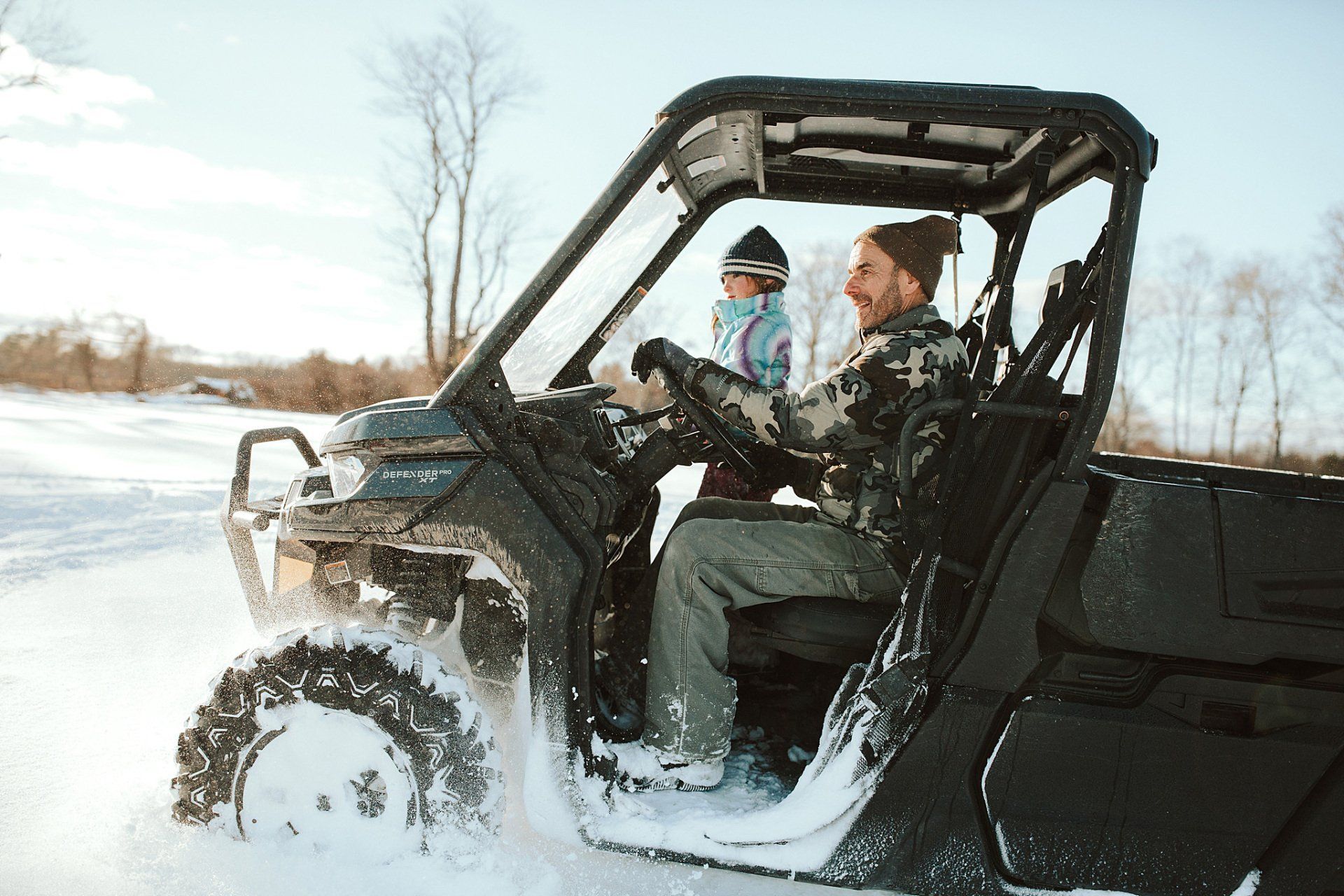Man driving an ATV in snow, with a child passenger. Snowy field and trees in the background, sunny day.