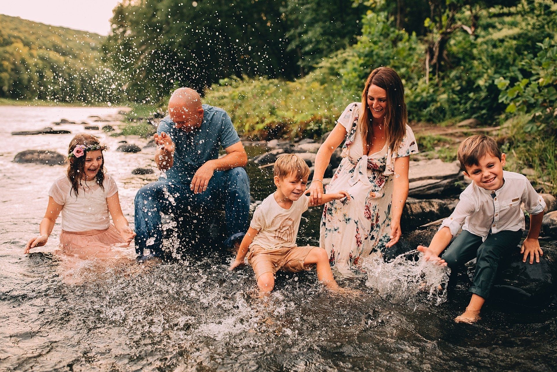 Family splashing in a river. Woman in floral dress, children smiling, water spraying.