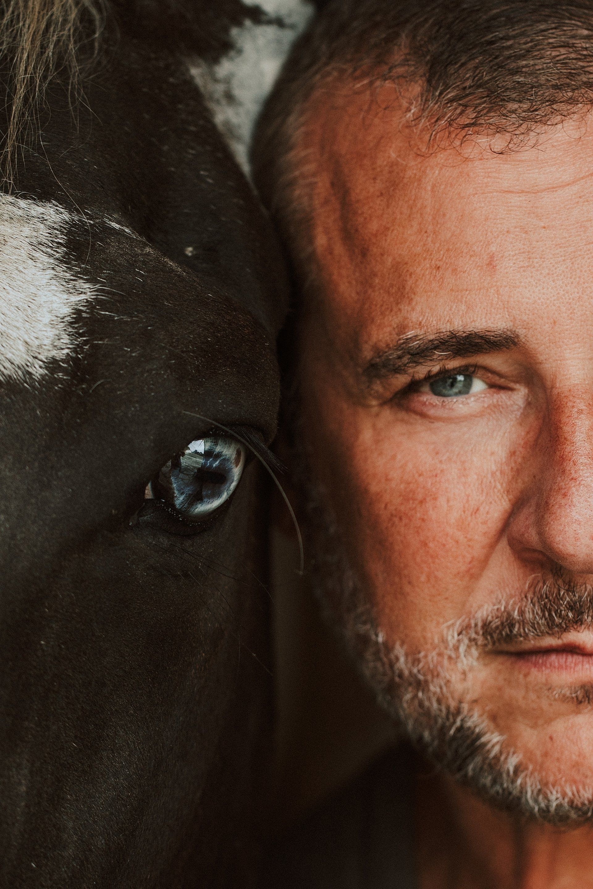 Man's face next to a dark horse's head; both looking toward the viewer.