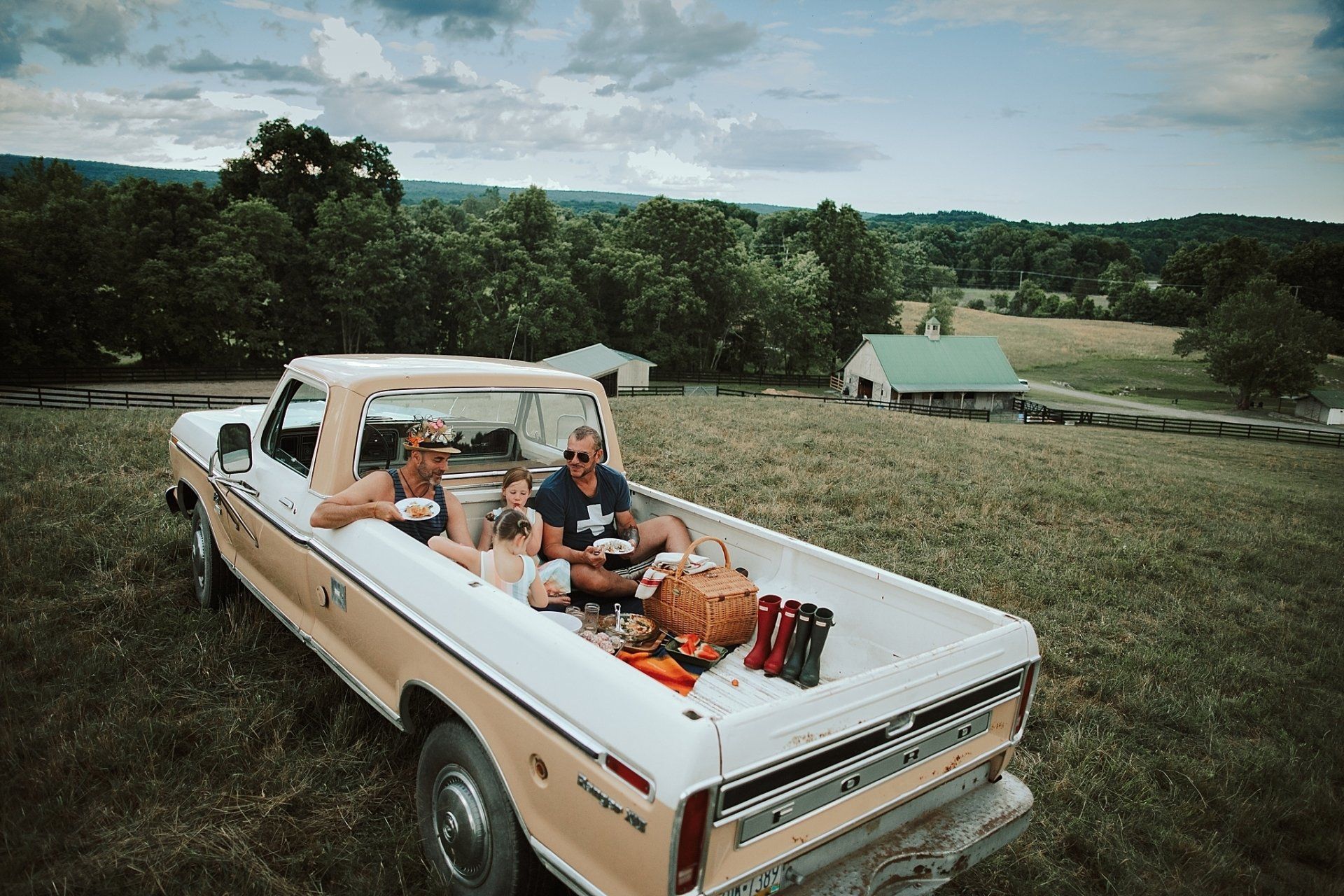 People having a picnic in the bed of a classic tan and white pickup truck, in a field with trees and a barn.