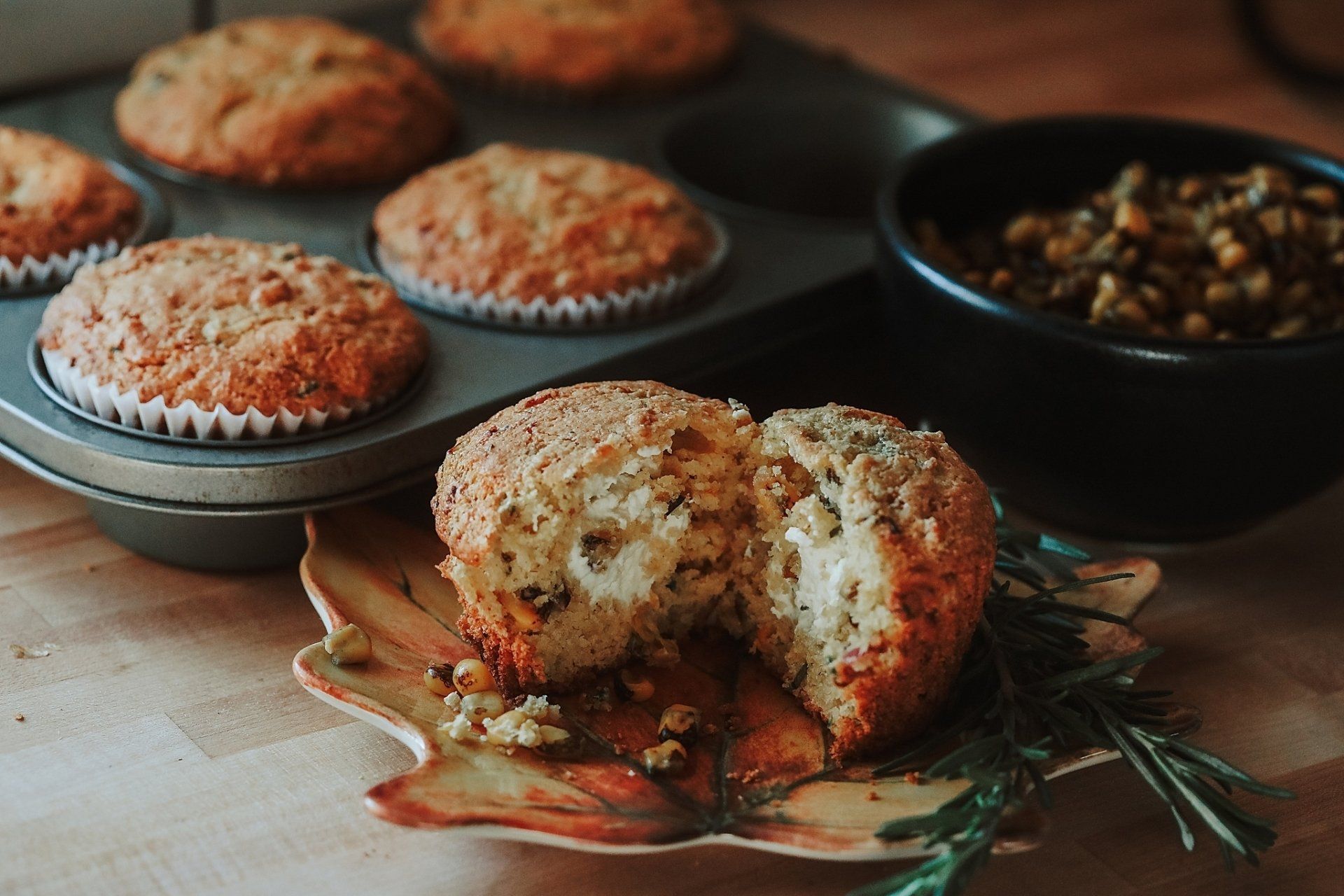 Muffins in a tin, one cut open on a leaf plate, with a bowl of food and rosemary sprig.