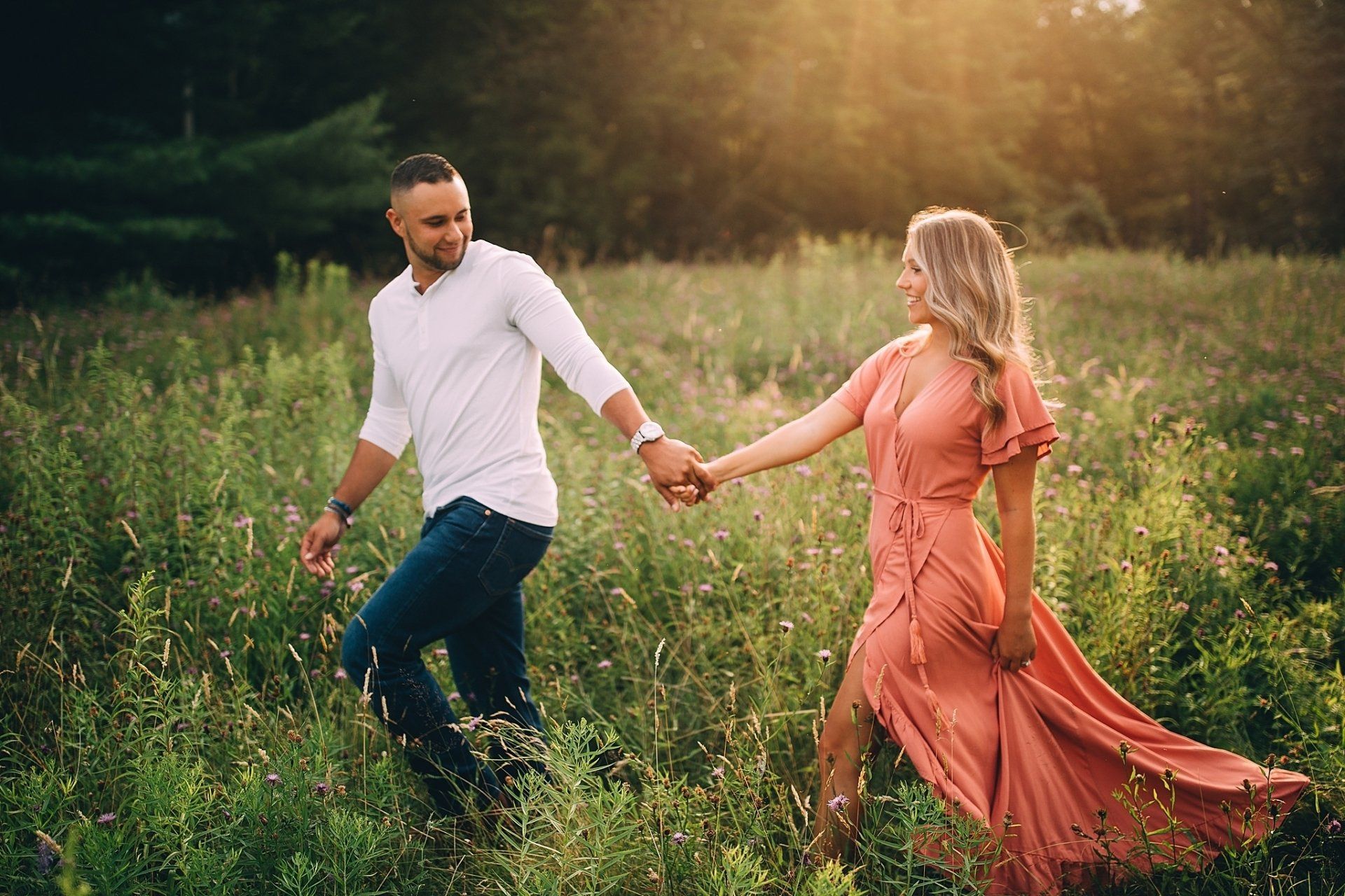 Couple walking hand-in-hand through tall grass at sunset. Woman in coral dress, man in white shirt and jeans. Hudson Valley photographer