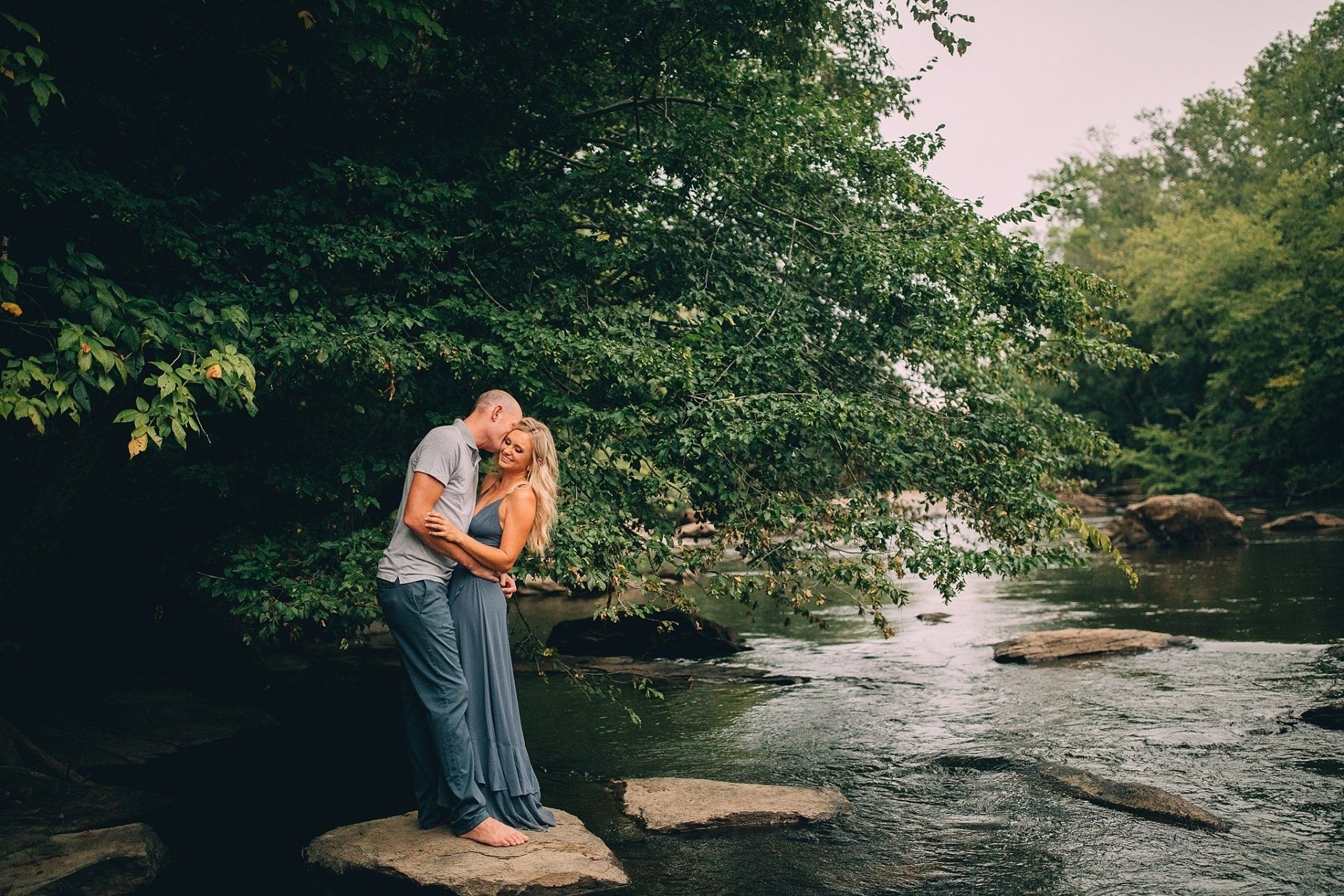 Couple embracing by a creek, under a large tree. The woman smiles, man kisses her cheek. Outdoors, daytime.