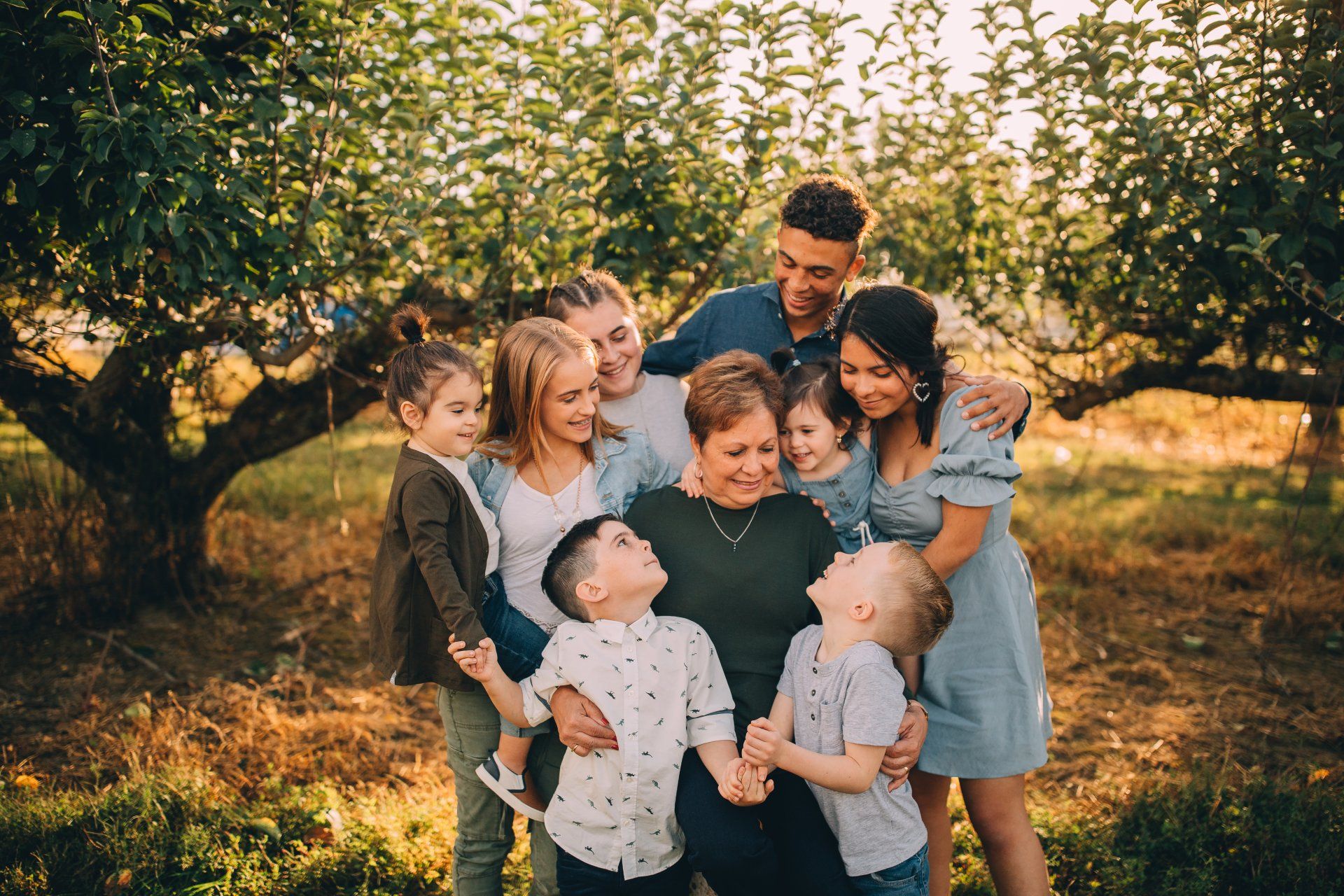 Family of nine embraces outdoors, smiles, under trees in autumnal light.