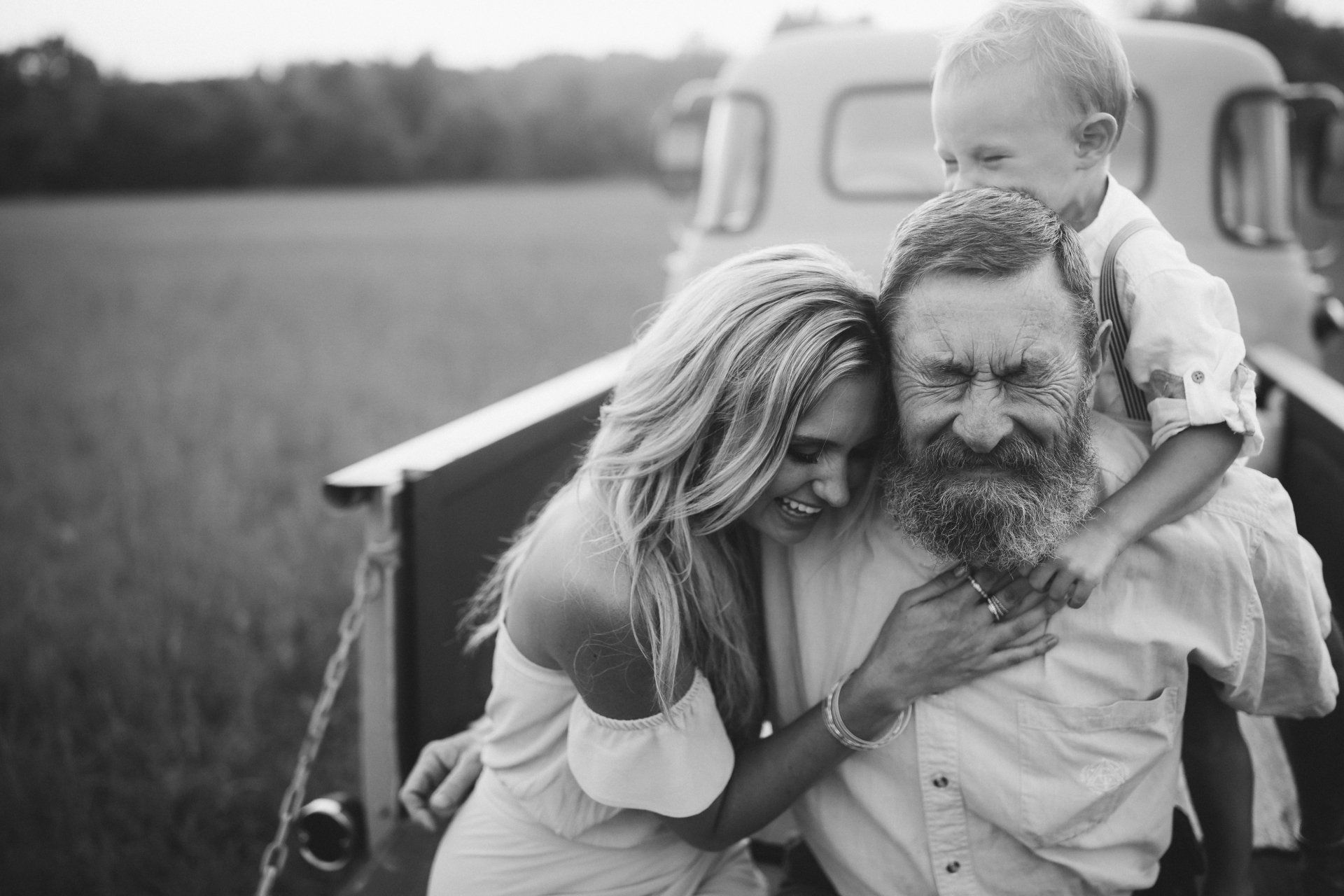 Family of three in a truck bed; woman laughing, man with child on shoulders, field background.