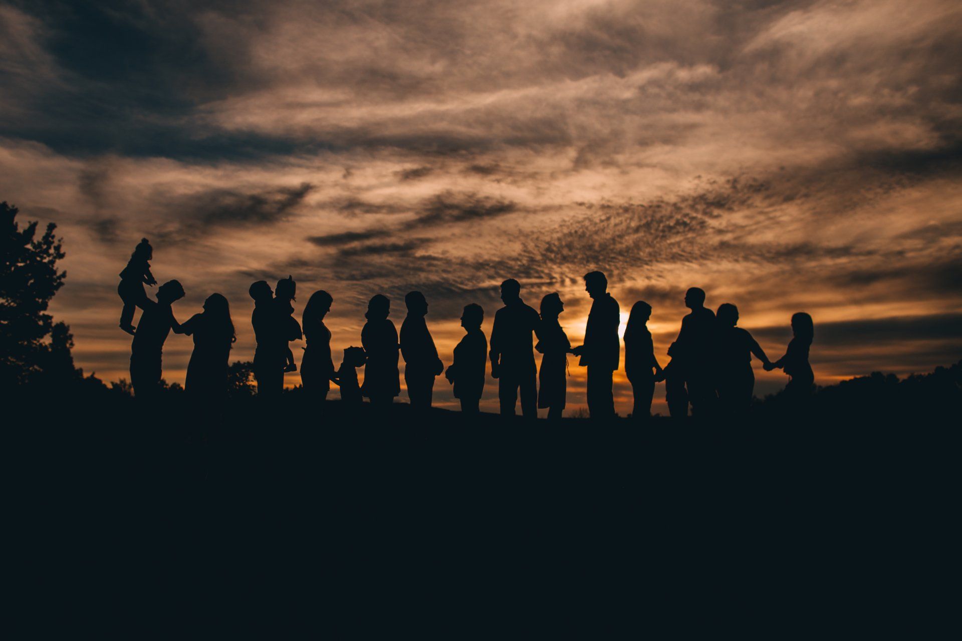 Silhouetted group of people against a sunset. Holding hands, some with children. Dark sky with clouds.