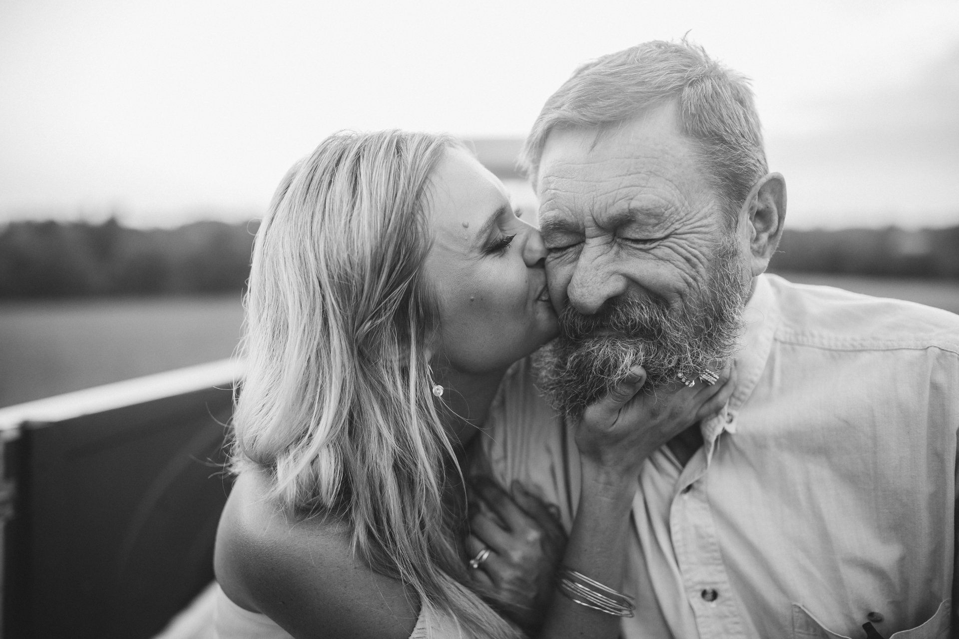 Woman kissing a bearded man's cheek, both smiling. Outdoors, soft light.