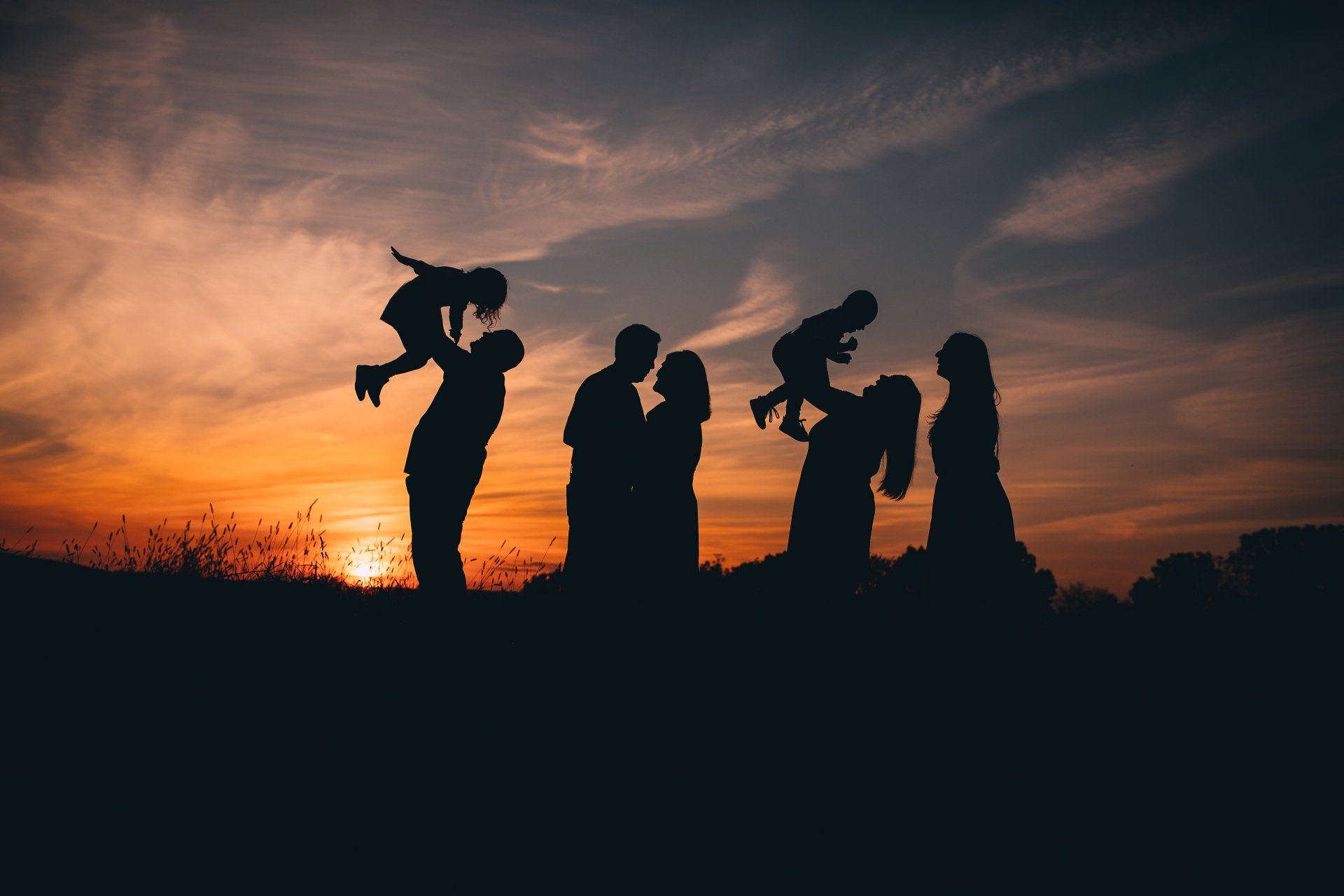 Silhouetted family at sunset: parents lifting children, embracing.