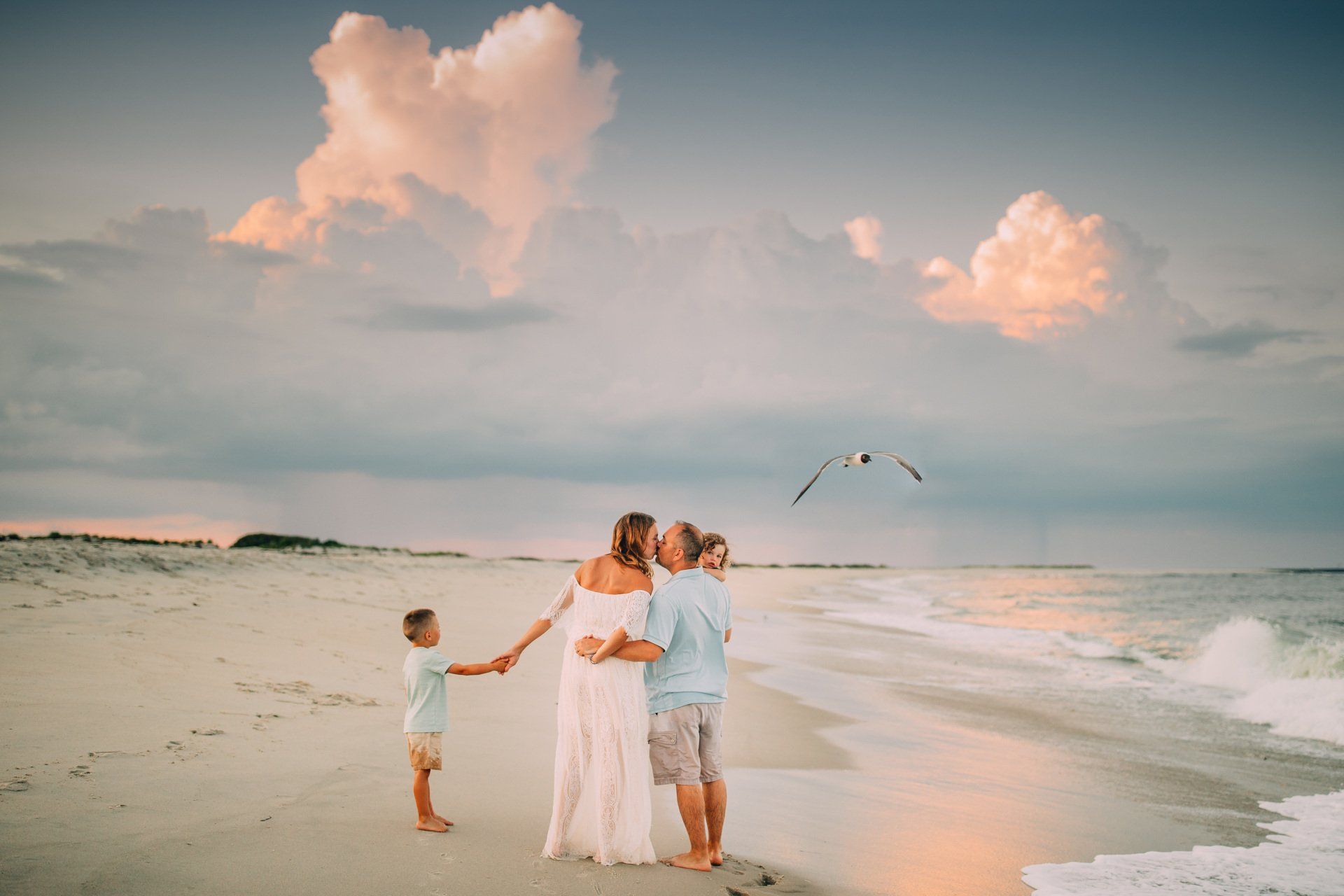 Family on beach at sunset, kissing, holding hands. Seagull flies overhead.