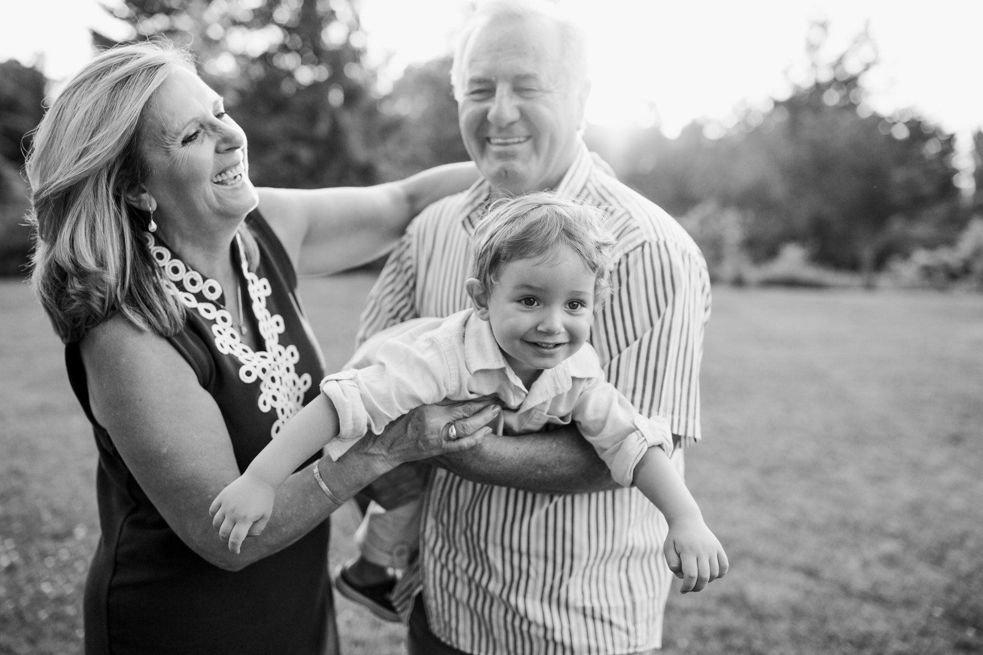 Woman and man holding a smiling toddler outdoors; both adults laugh, sunlight in background.