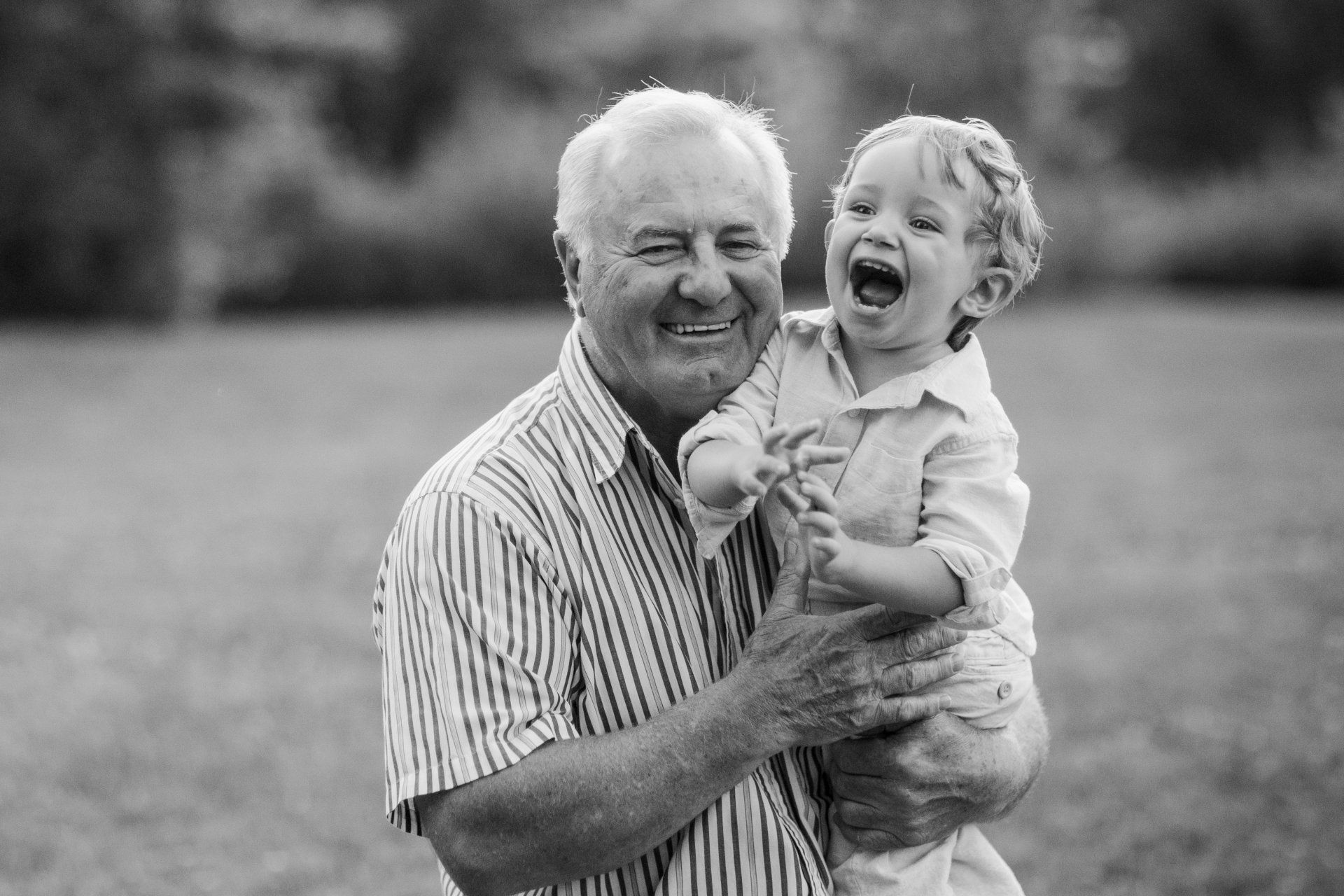 A smiling older man holds a laughing child outdoors; both are happy.