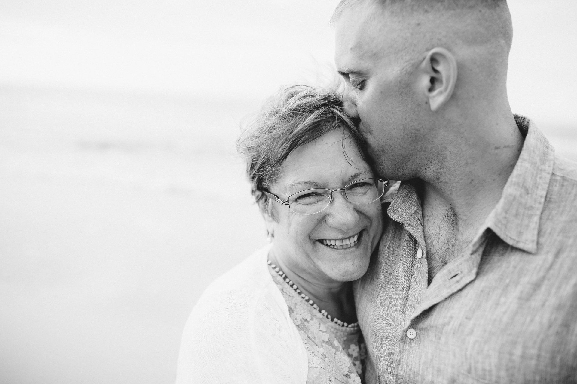 Man kisses woman's forehead; they smile at the beach, both in light-colored clothing.