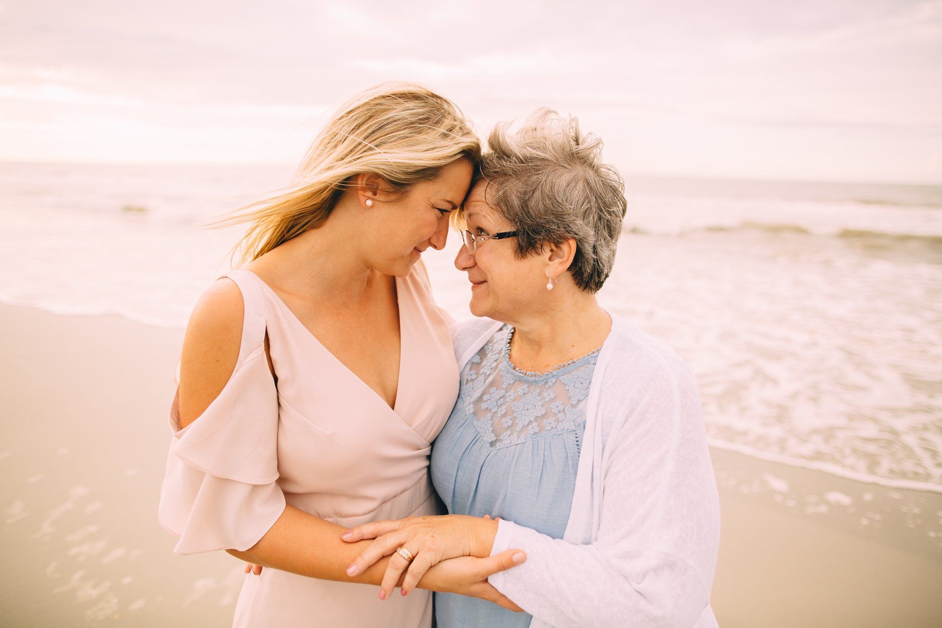 Woman in pink dress embraces older woman on a beach, heads touching, smiles.