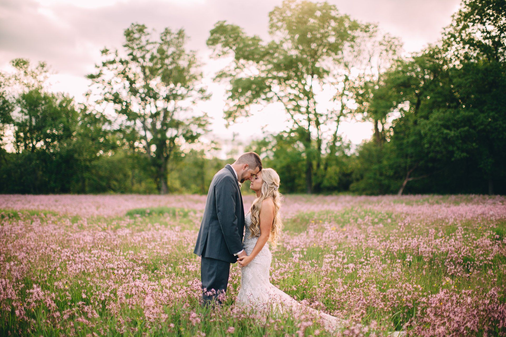 Bride and groom in a field of pink flowers, heads touching, holding hands. Trees in the background.